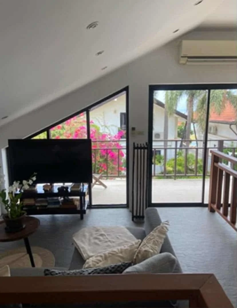 Living room with a sofa, a potted plant, a side table, a TV on a stand, a balcony with pink flowers, and sliding glass doors leading outside.