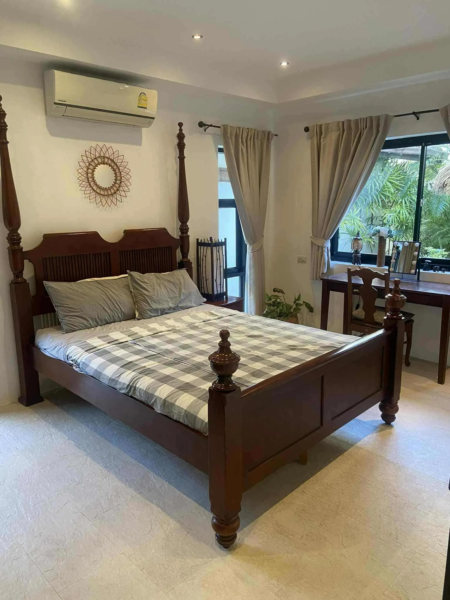A bedroom with a wooden four-poster bed, checkered bedsheet, two pillows, side table, and a desk near the window with curtains, plants, and decorative items.