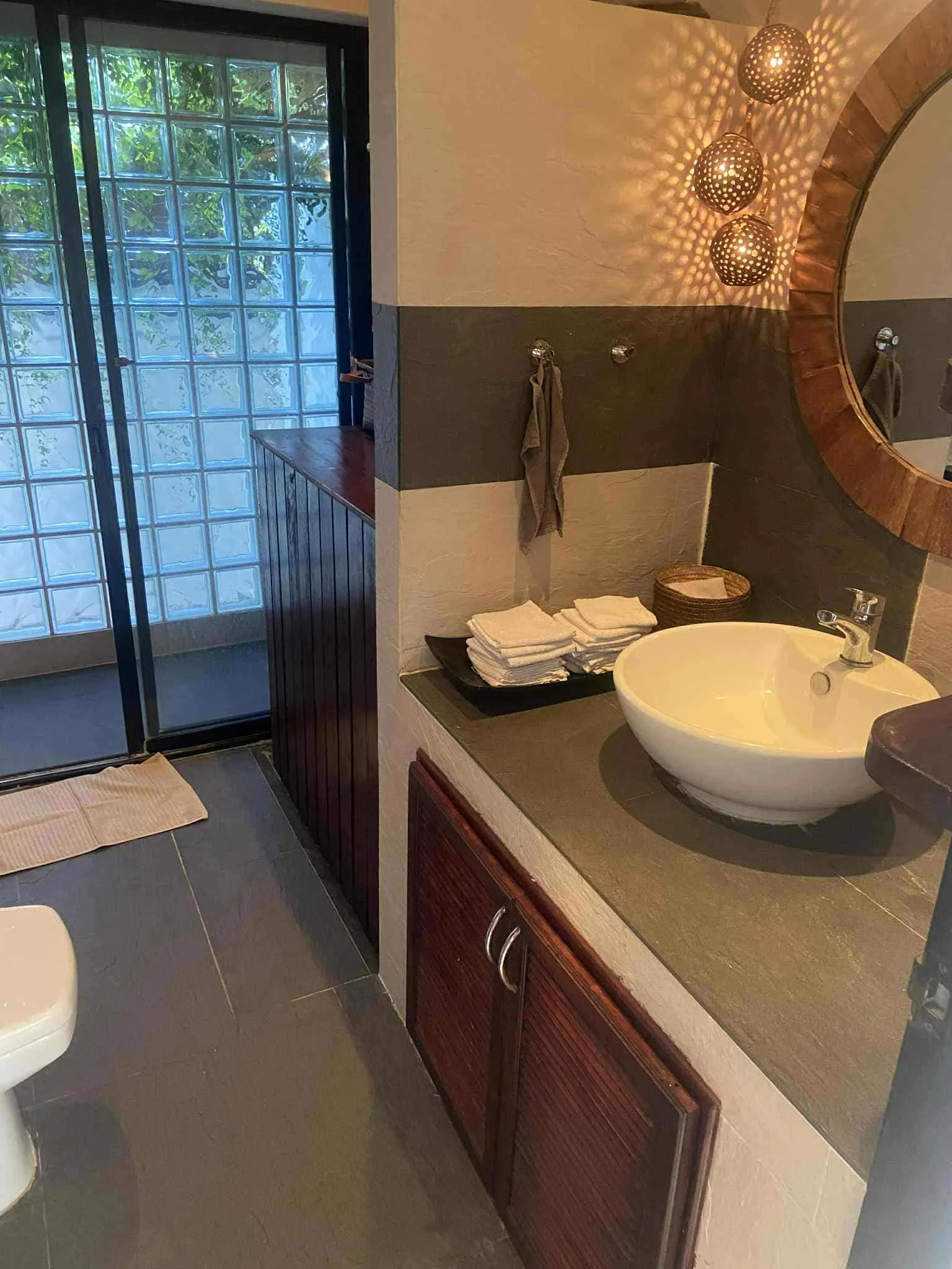 A bathroom with a white vessel sink on a black countertop, a round mirror with a wooden frame, dark wooden cabinets, folded towels, a small basket, hanging towels, and a decorative light fixture casting patterned shadows, with a glass block wall and 