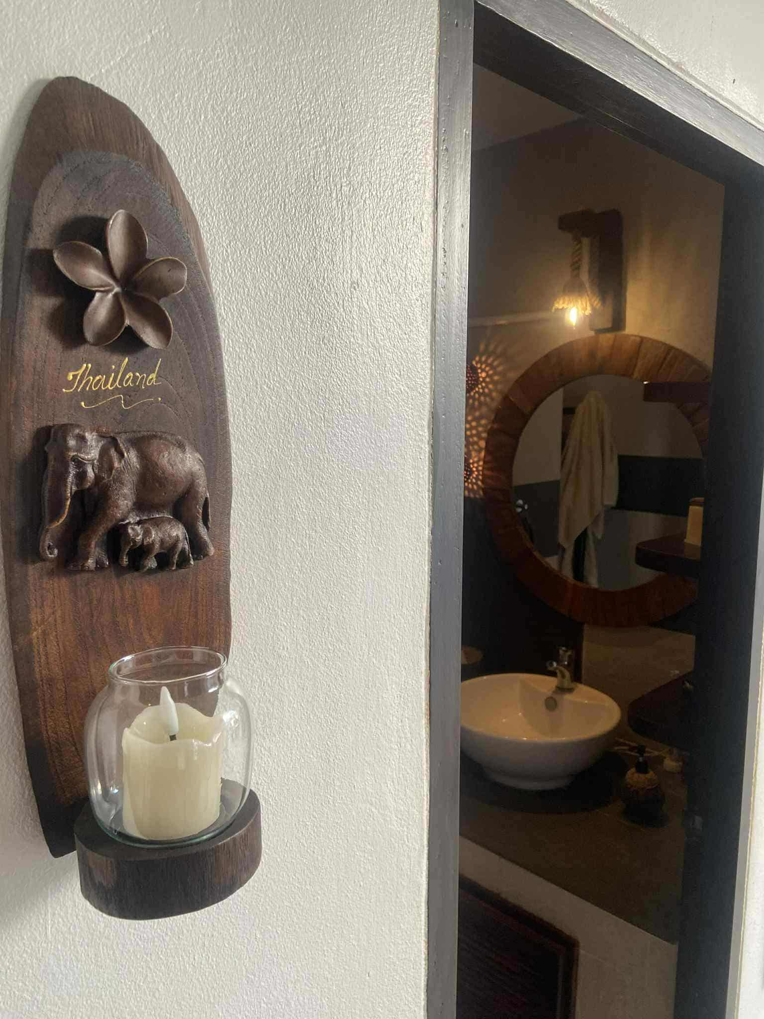 Decorative wall plaque with the word 'Thailand', a wooden elephant sculpture, and a glass candle holder with a white candle, hung on a textured wall near a doorway leading to a bathroom with a round mirror and a sink.