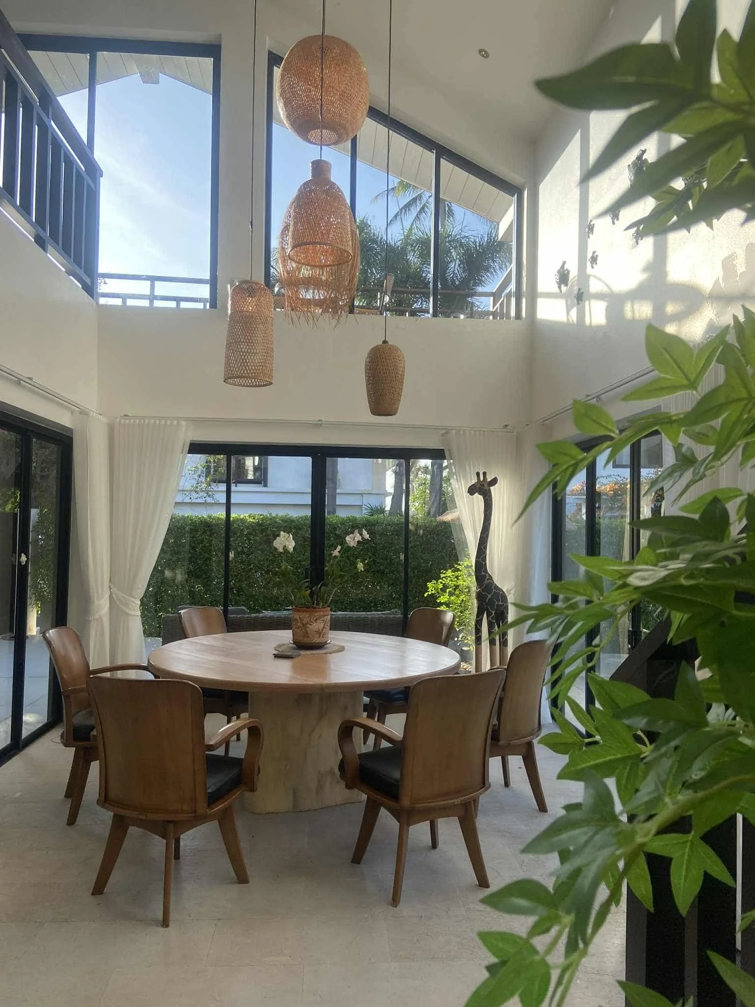 Bright dining room with large windows, round wooden table, six wooden chairs, hanging woven light fixtures, potted orchid, large plant, and a giraffe sculpture in the corner.
