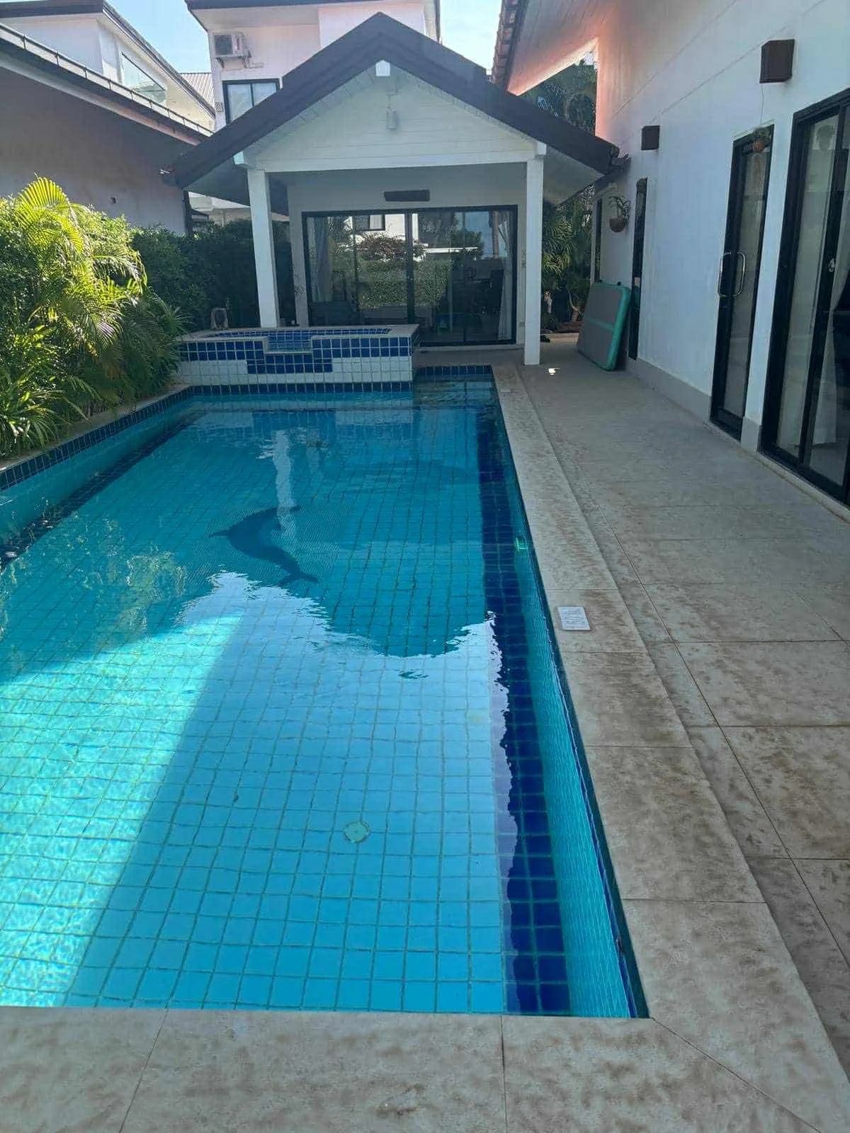 A backyard swimming pool with clear blue water, surrounded by beige stone tiles, next to a white house with glass sliding doors. There is a small covered patio area with outdoor furniture and lush green plants lining the fence on the left side.