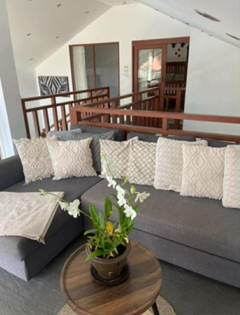 Living room with sectional sofa, decorative pillows, a round wooden table with a potted plant, and a wooden railing overlooking a loft area with a door and artwork on the wall.