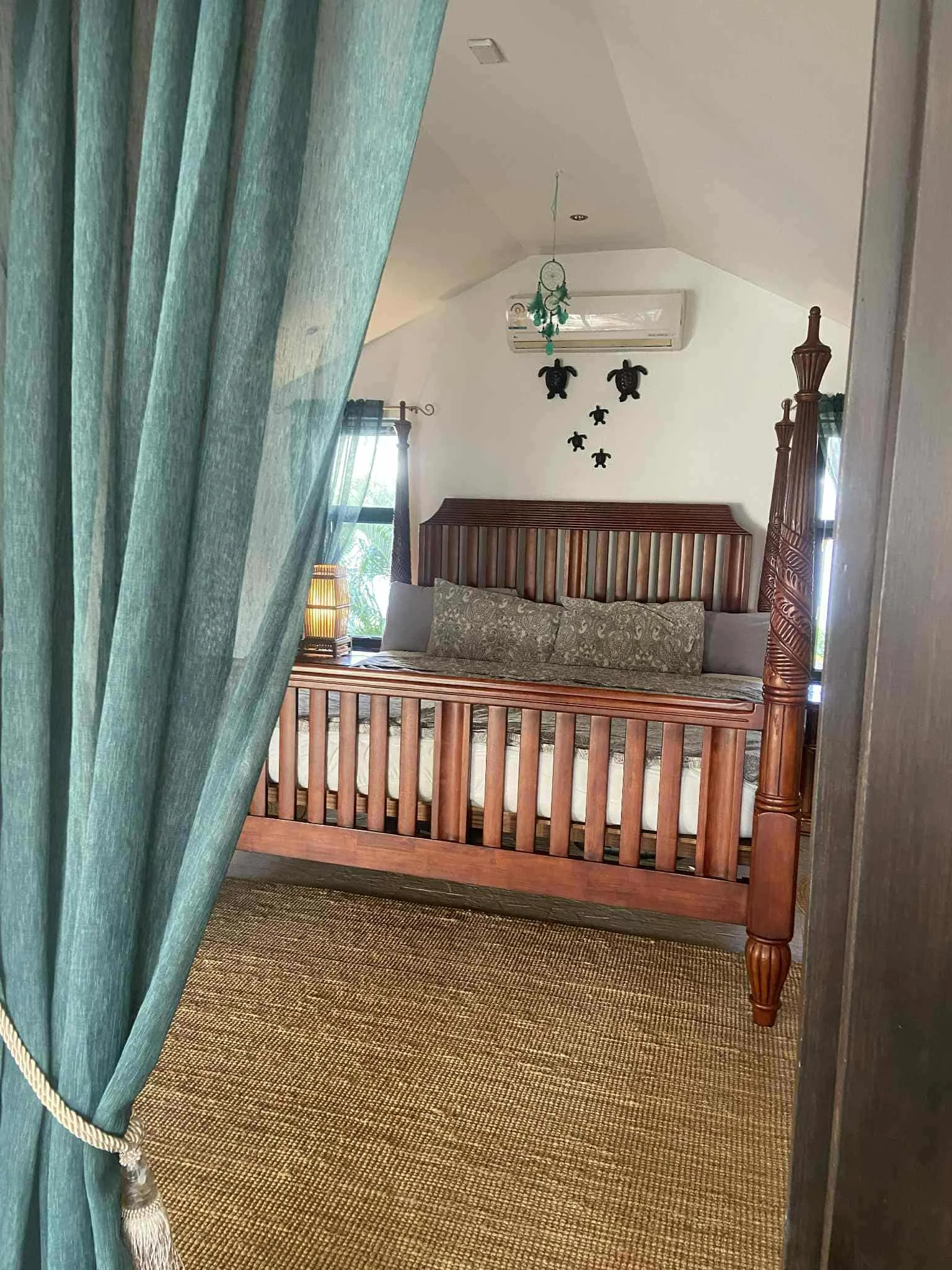 View of a bedroom with a wooden bed, bedside table, lamp, window with curtains, wall decorations of teddy bears, and a ceiling air conditioning unit.