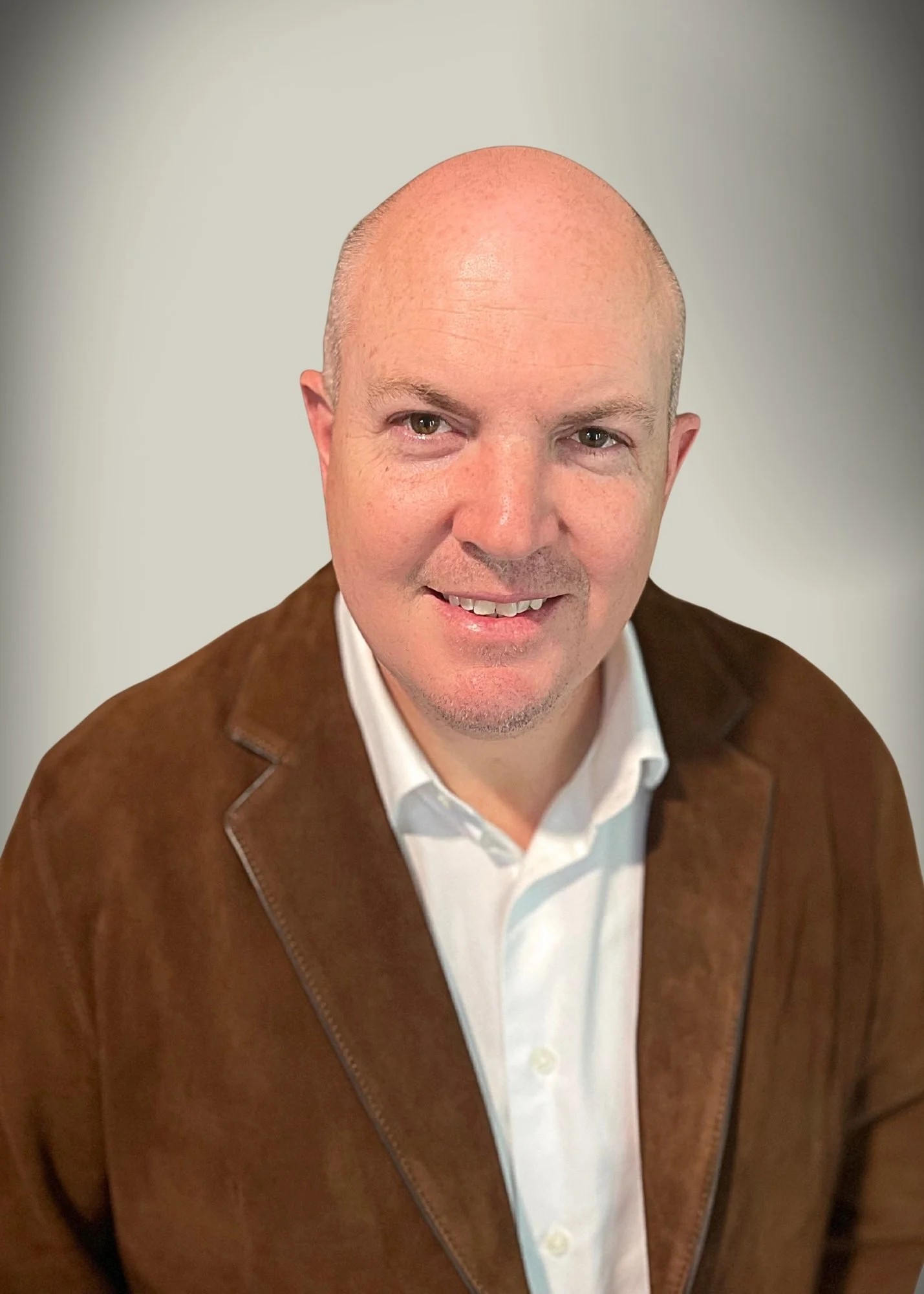 A bald man smiling, wearing a white shirt and a brown jacket, against a neutral background.