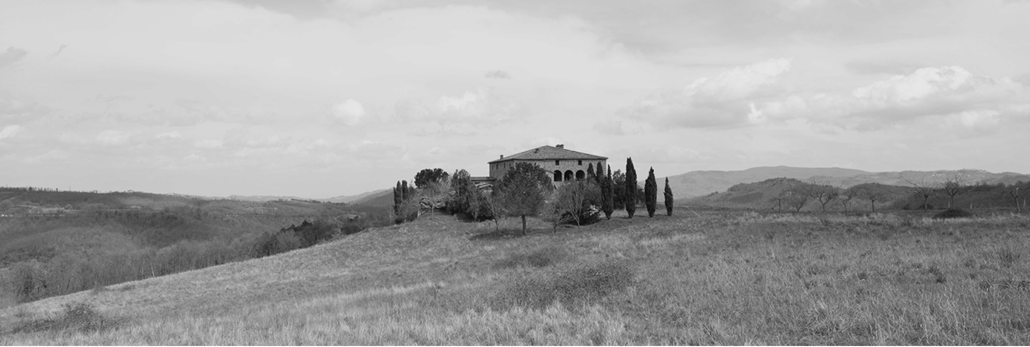 A house on a hill surrounded by trees in a rural landscape with rolling hills in the background, black and white photograph.