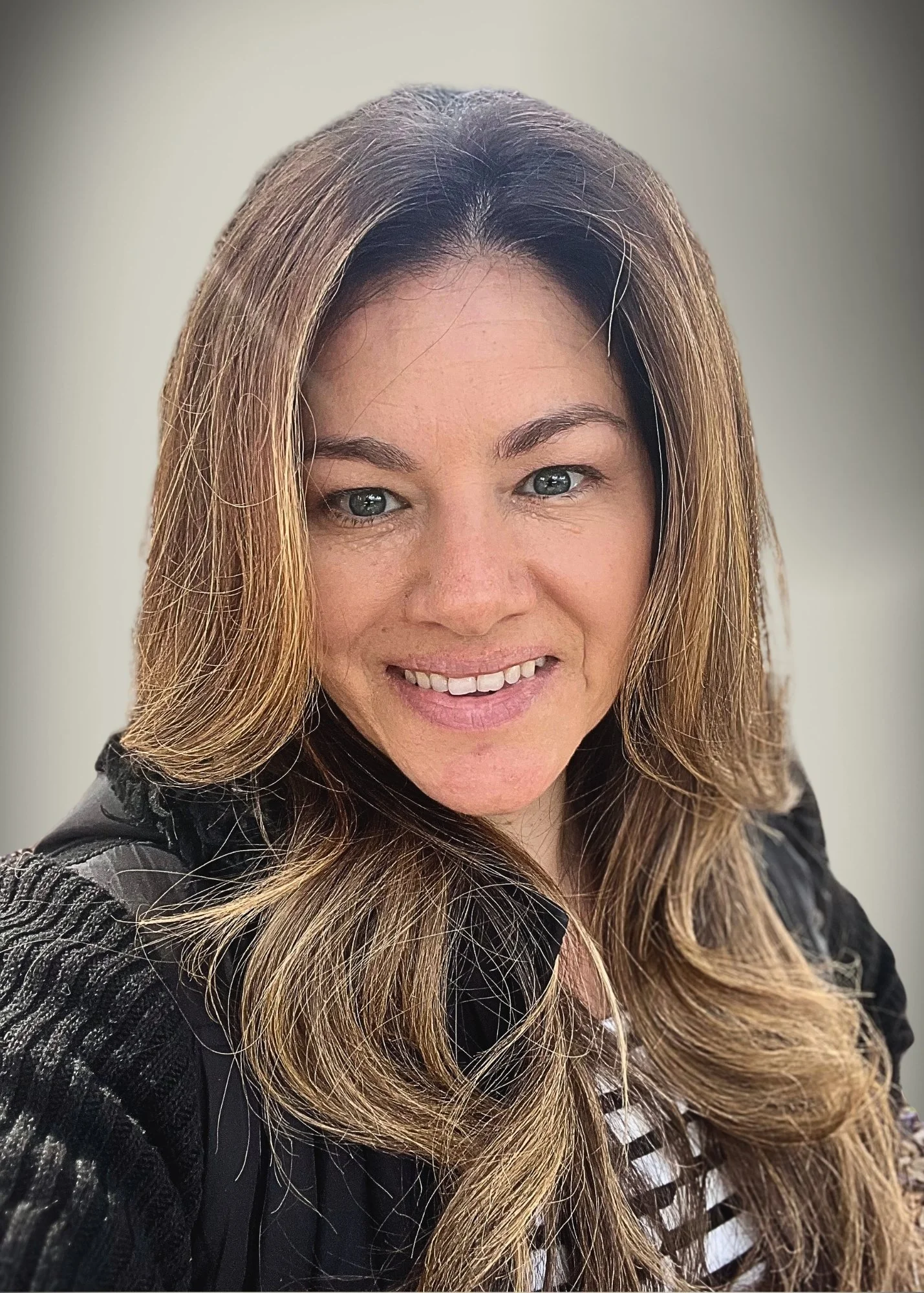 A woman with long, wavy brown hair and light skin, smiling at the camera, wearing a black jacket and a striped shirt.