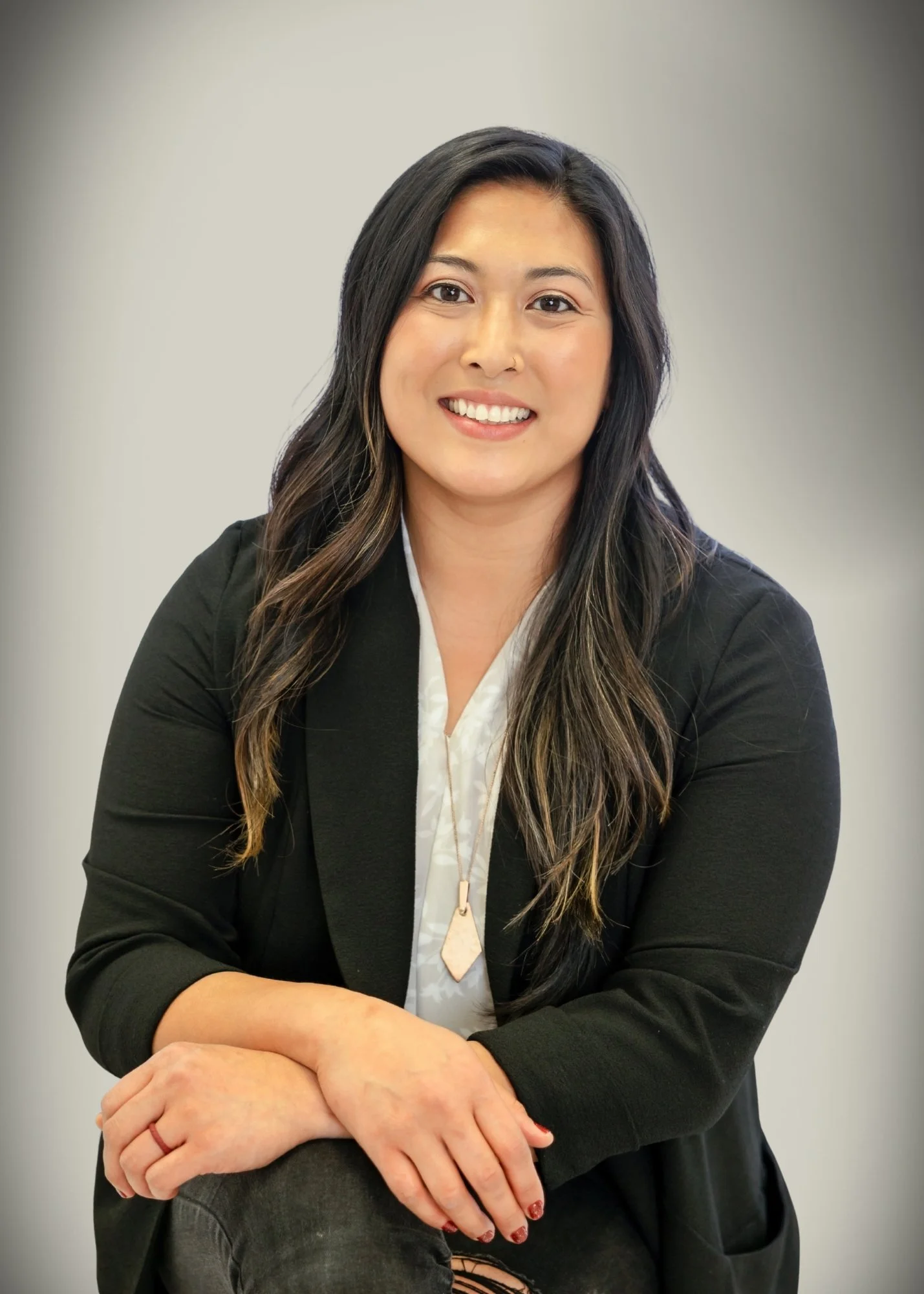A woman with long dark hair, wearing a black blazer over a white blouse, posed sitting with her arms crossed on her knee, smiling at the camera against a plain background.