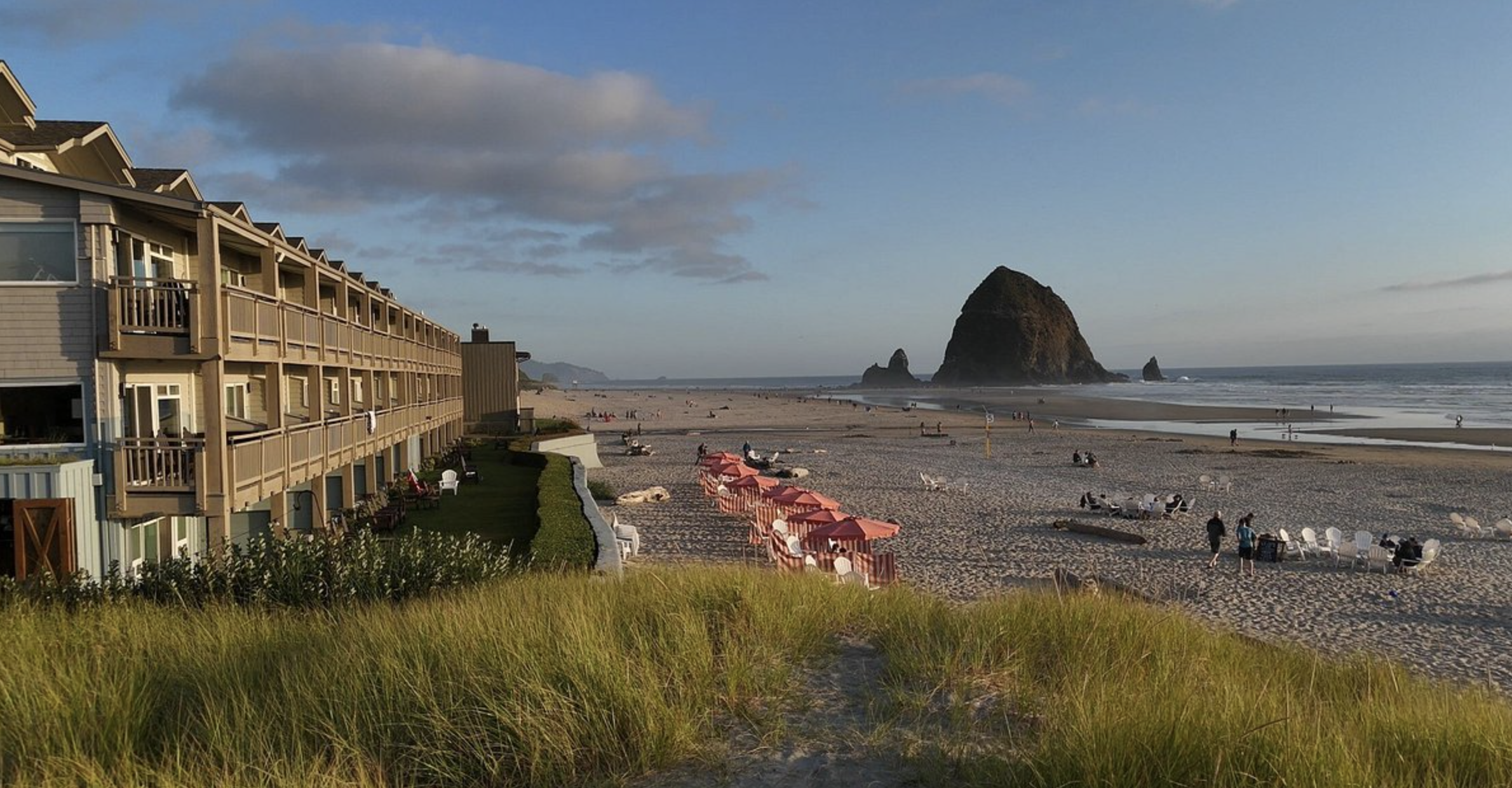 Beach with sand dunes and grass in the foreground, multi-story beachside hotel on the left, large sea stack in the distance, and people relaxing on the beach under umbrellas and walking along the shoreline.
