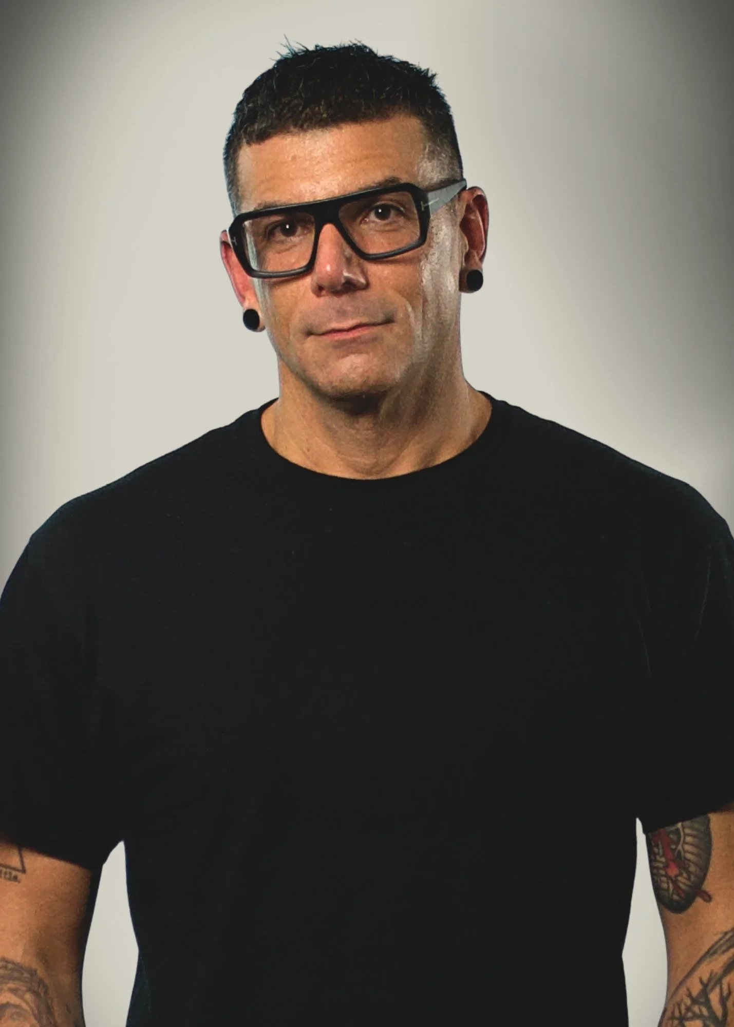 Portrait of a man with short dark hair, wearing large black-rimmed glasses, black earrings, and a black t-shirt, standing against a neutral background.