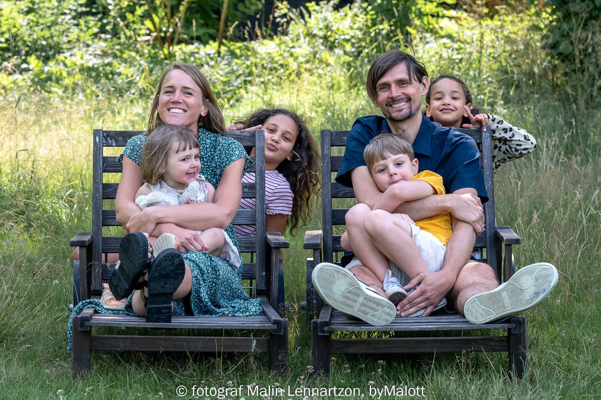 Porträtt av familj som sitter på trästolar i naturen och håller om varandra och är glada.