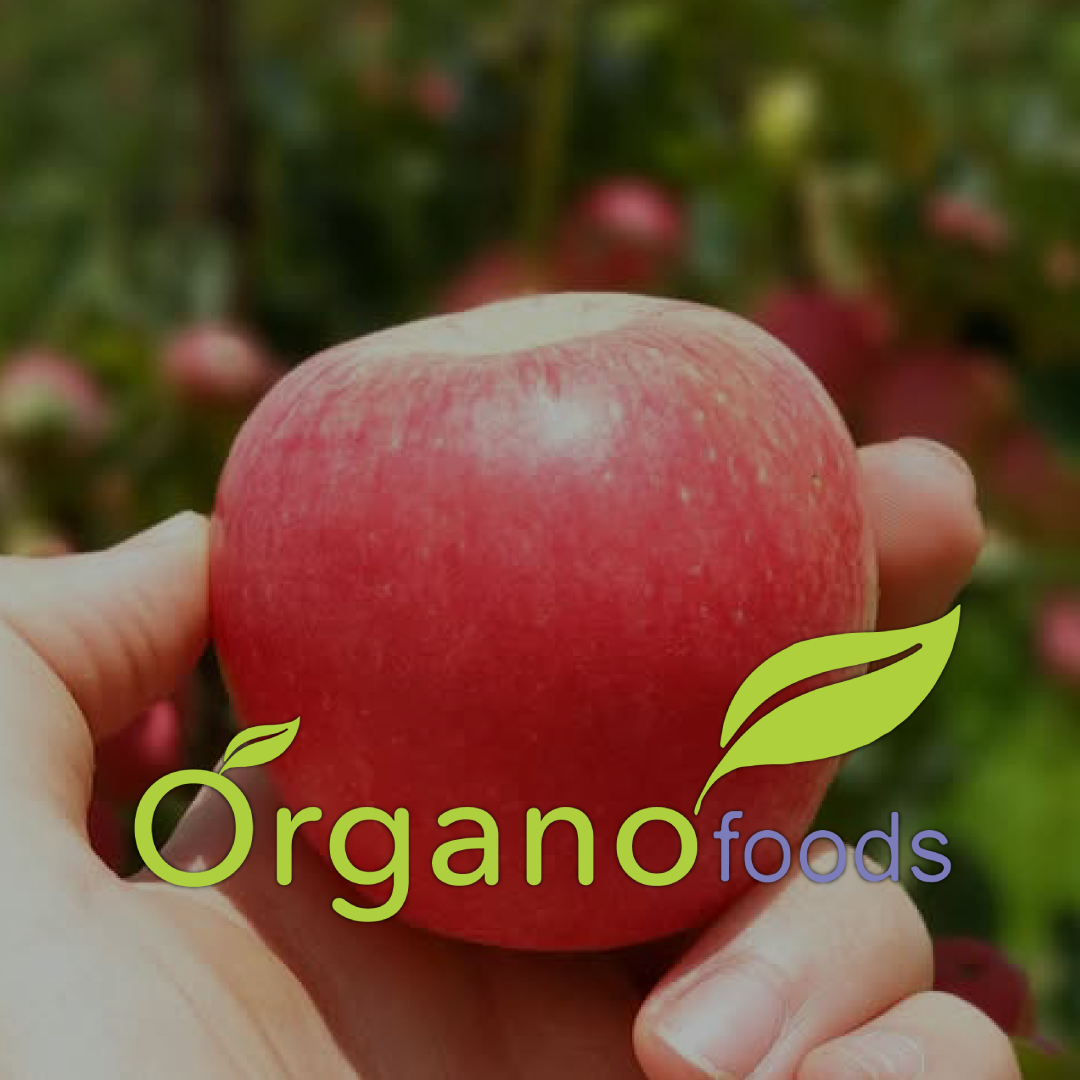 Close-up of a hand holding a red apple with a blurred background of apple trees, featuring a logo with green leaves and the text "Organofoods."