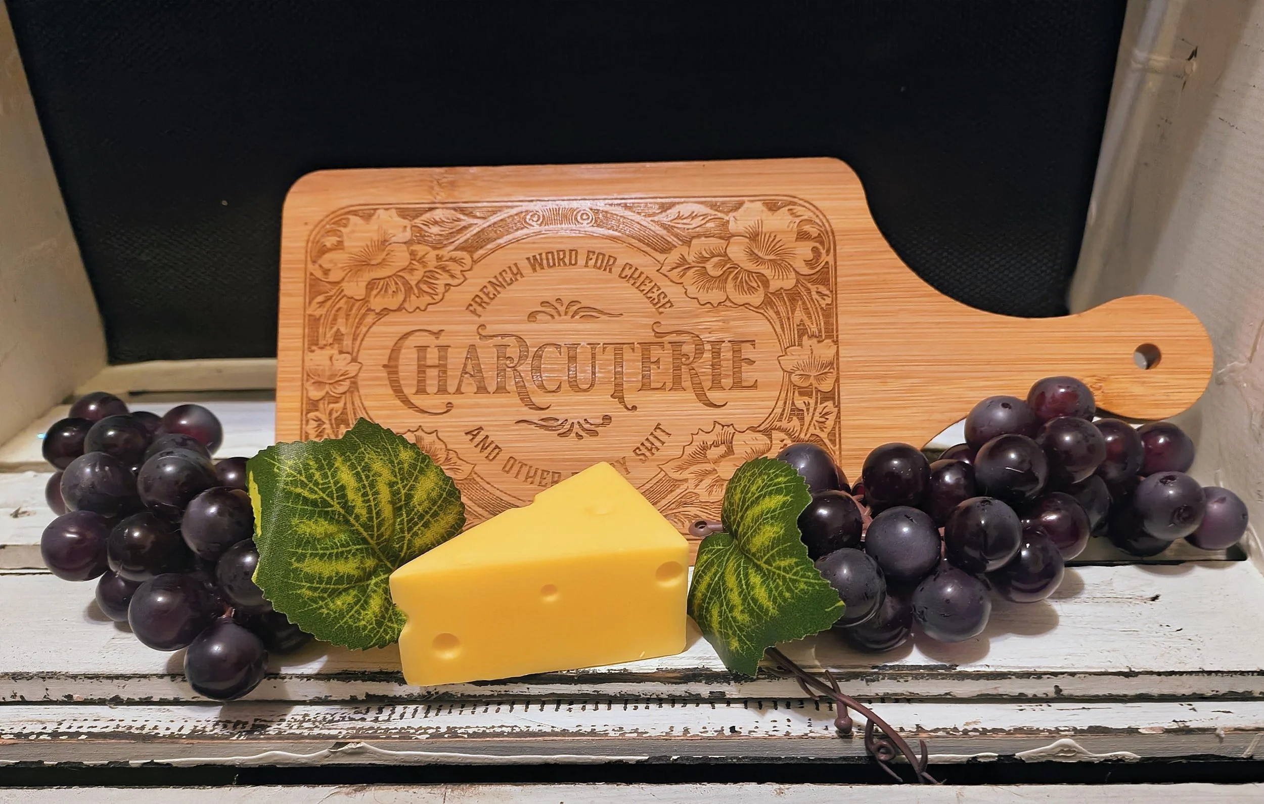 Grapes, a wedge of cheese with leaves, and a wooden cutting board with engraved text.