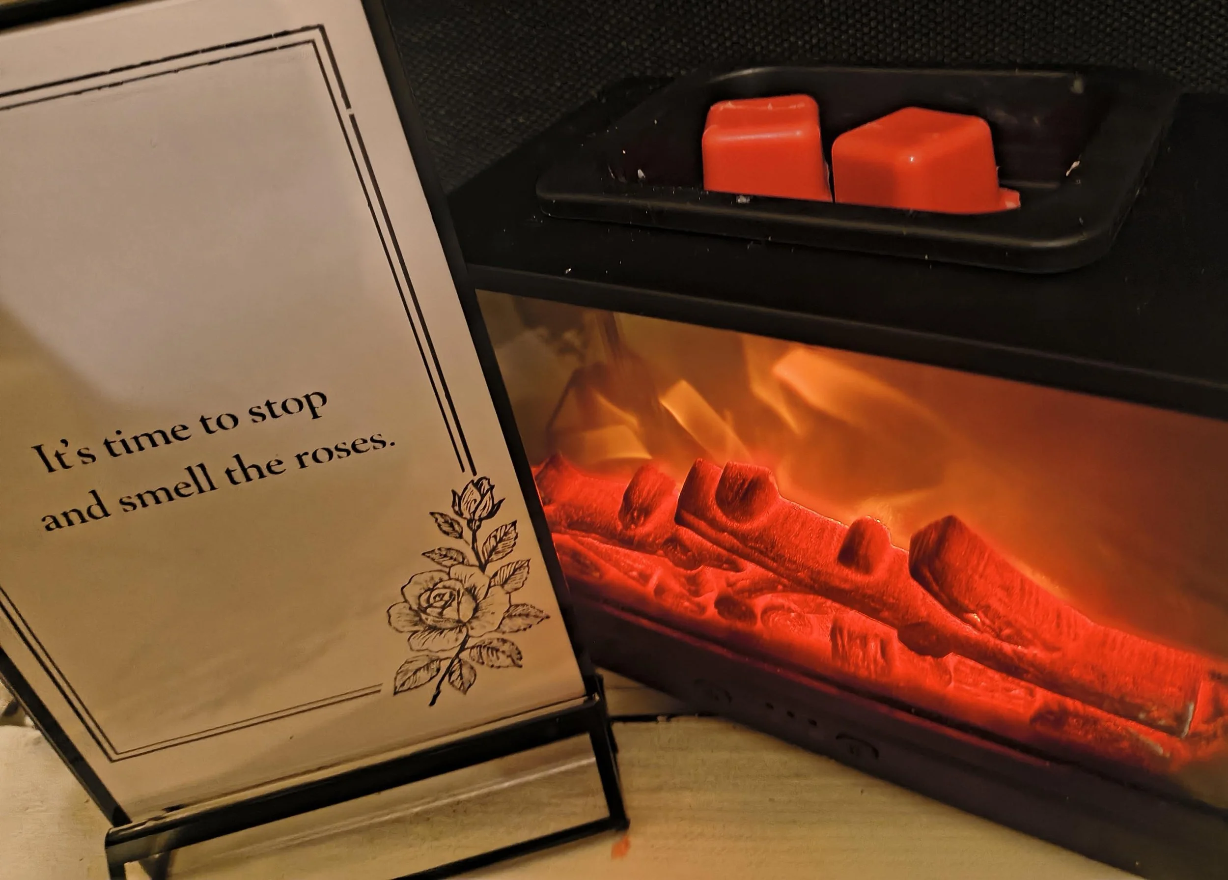 A sign with the text 'It's time to stop and smell the roses' and a rose illustration, placed in front of a fireplace with glowing red-orange logs.