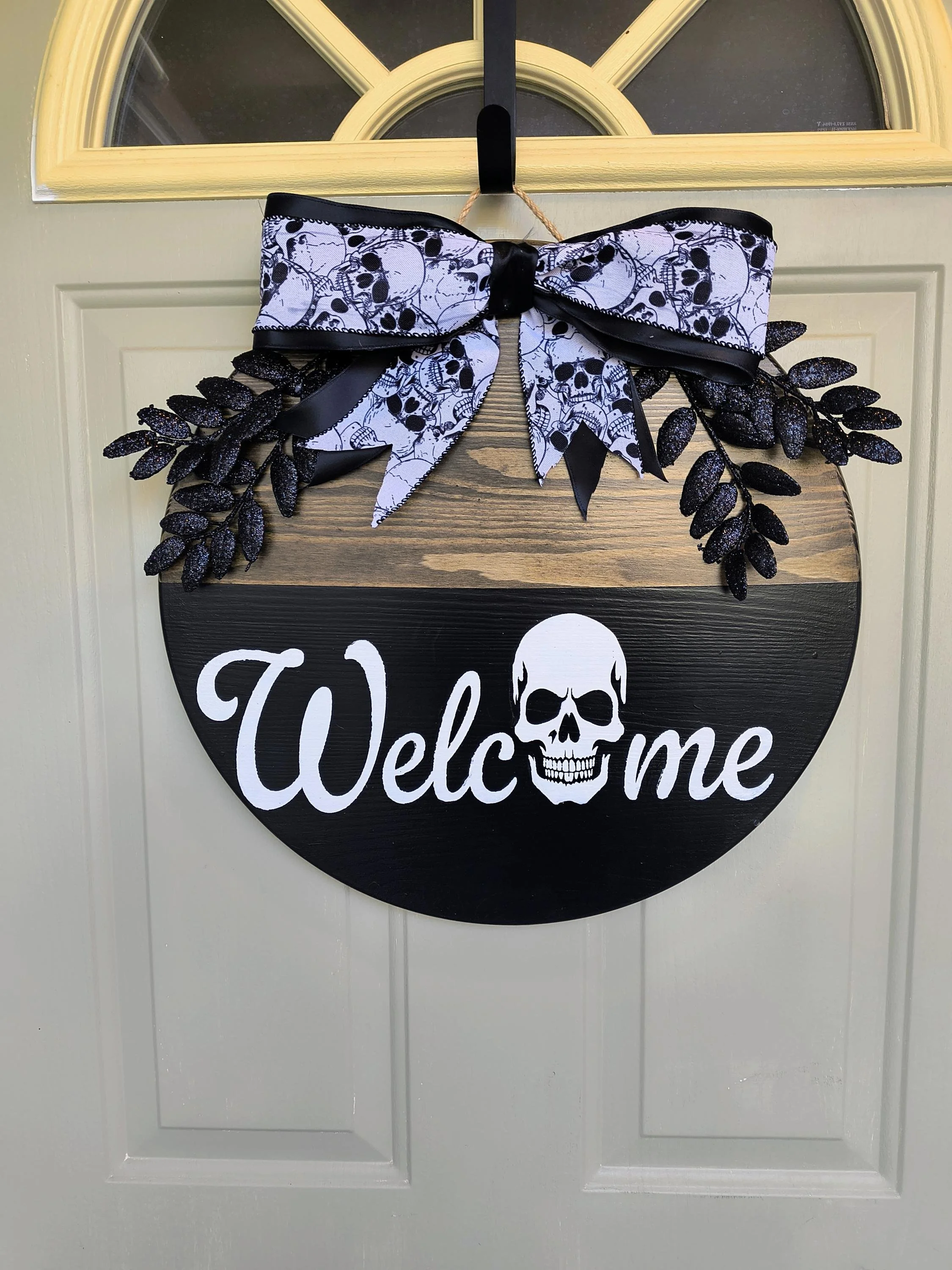 Decorative Halloween door sign with a black and wood-colored half circle background. It features a white skull, the word 'Welcome' in white cursive, a black ribbon with a pattern of white skulls and crossbones, and black glittery leaves, hanging on a