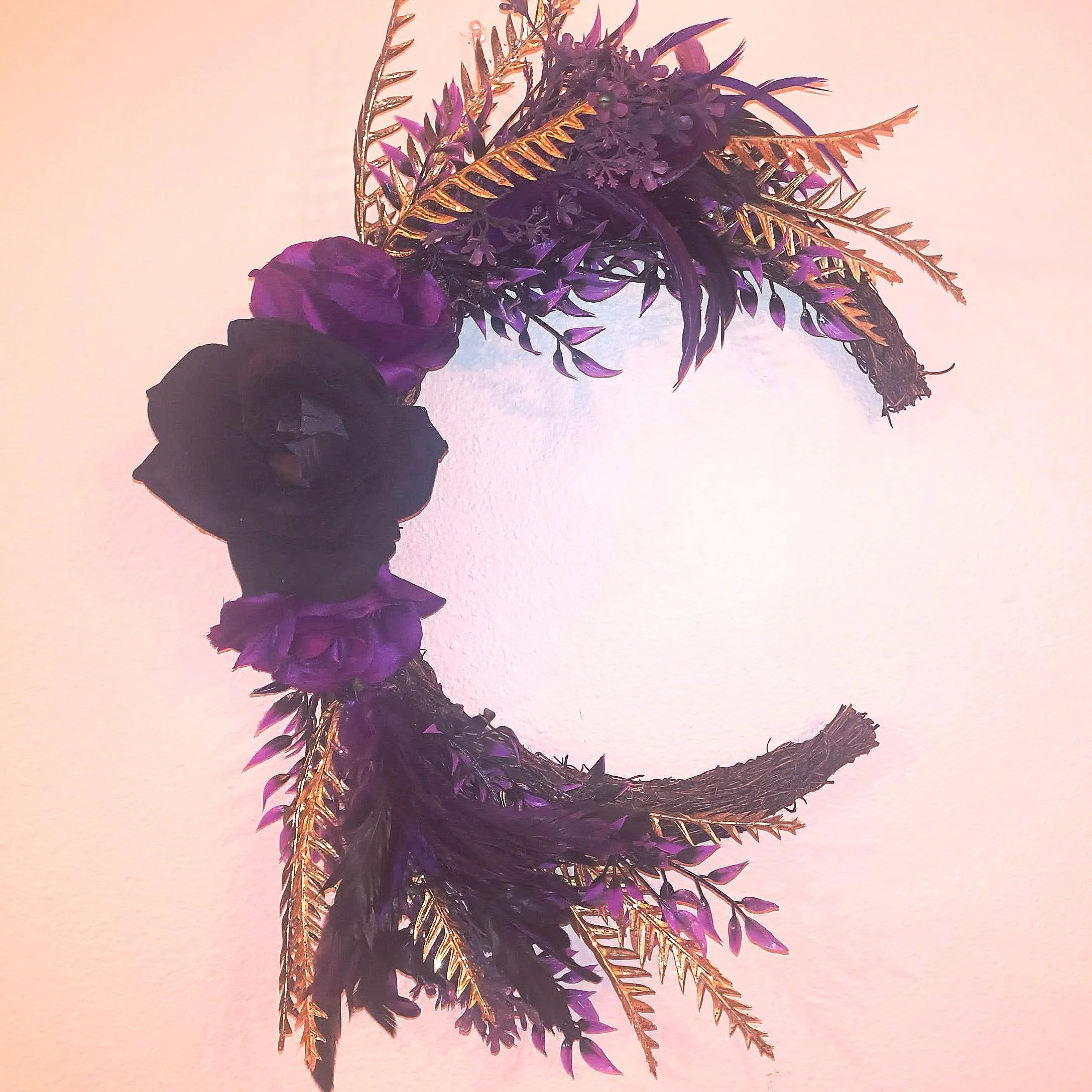 A decorative crescent-shaped wreath made of faux purple and black roses, gold-colored leaves, and black feathers, mounted on a plain light-colored wall.