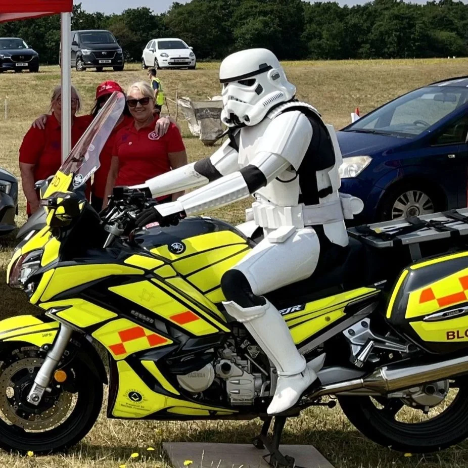 A person dressed as a Stormtrooper from Star Wars riding a yellow police motorcycle, with two smiling women in red shirts in the background.