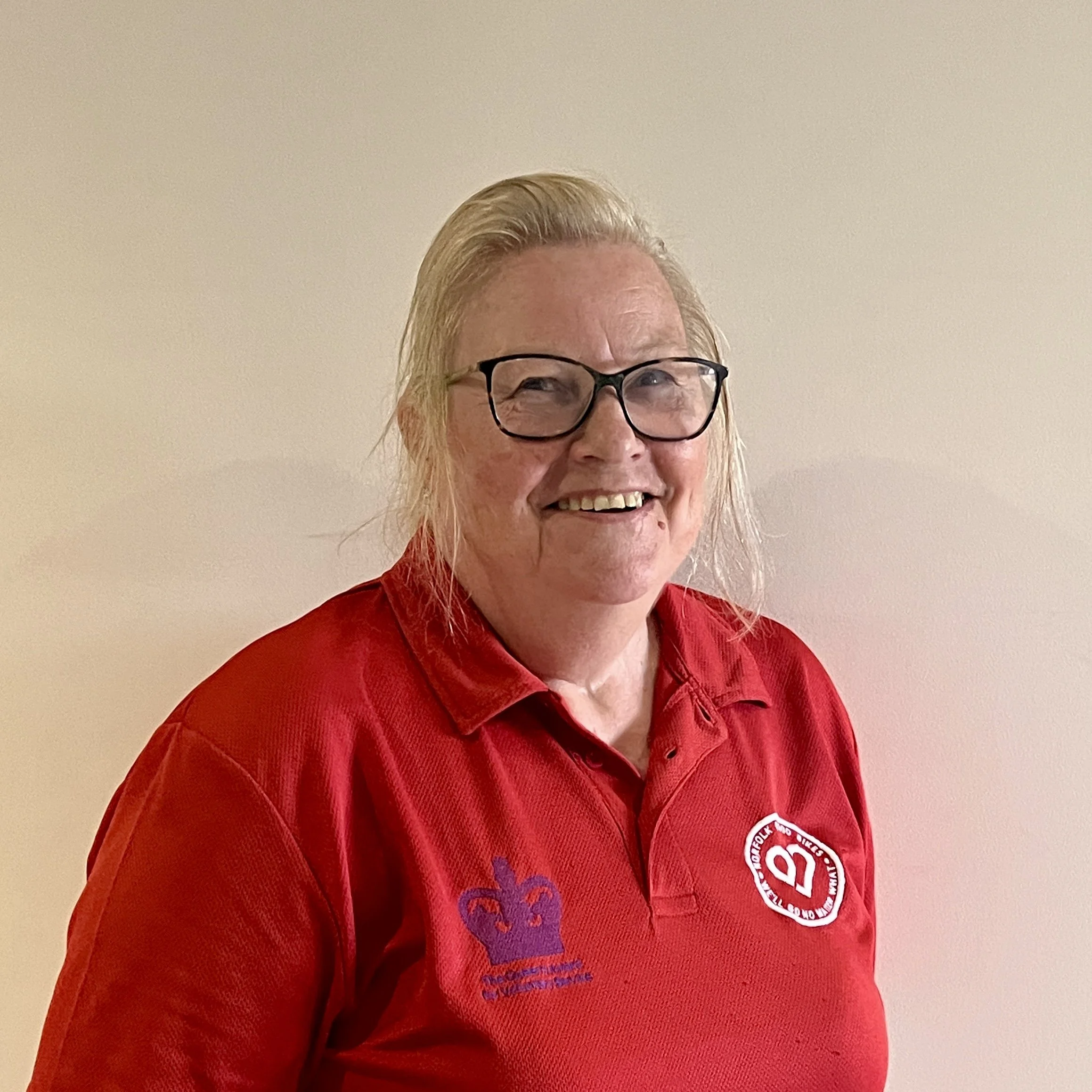 A female volunteer with blonde hair and glasses, wearing a blue floral shirt, outdoors near a body of water with trees and houses in the background.