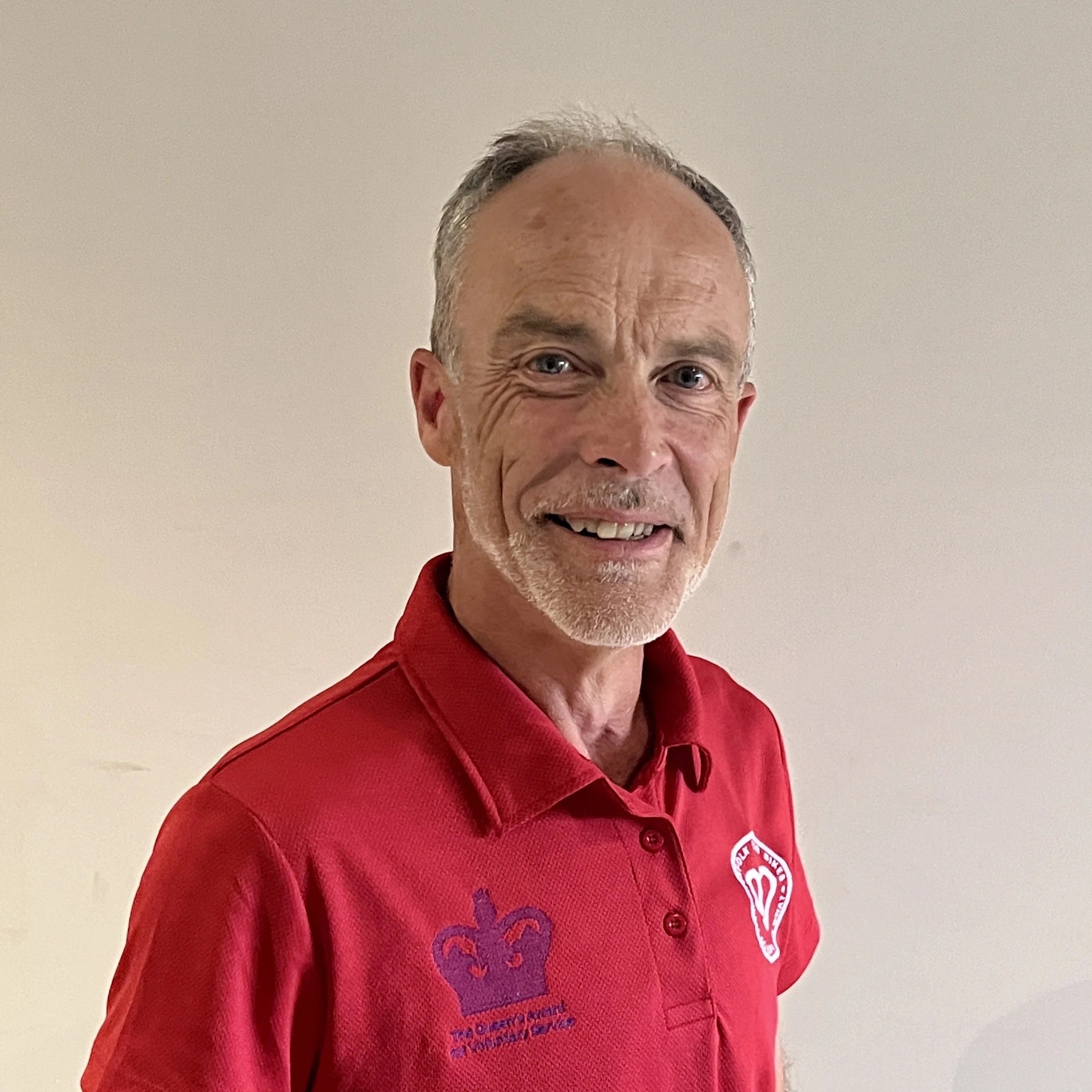 A smiling middle-aged man with gray hair and a beard, wearing a red polo shirt with logos on the chest, standing against a plain beige wall.