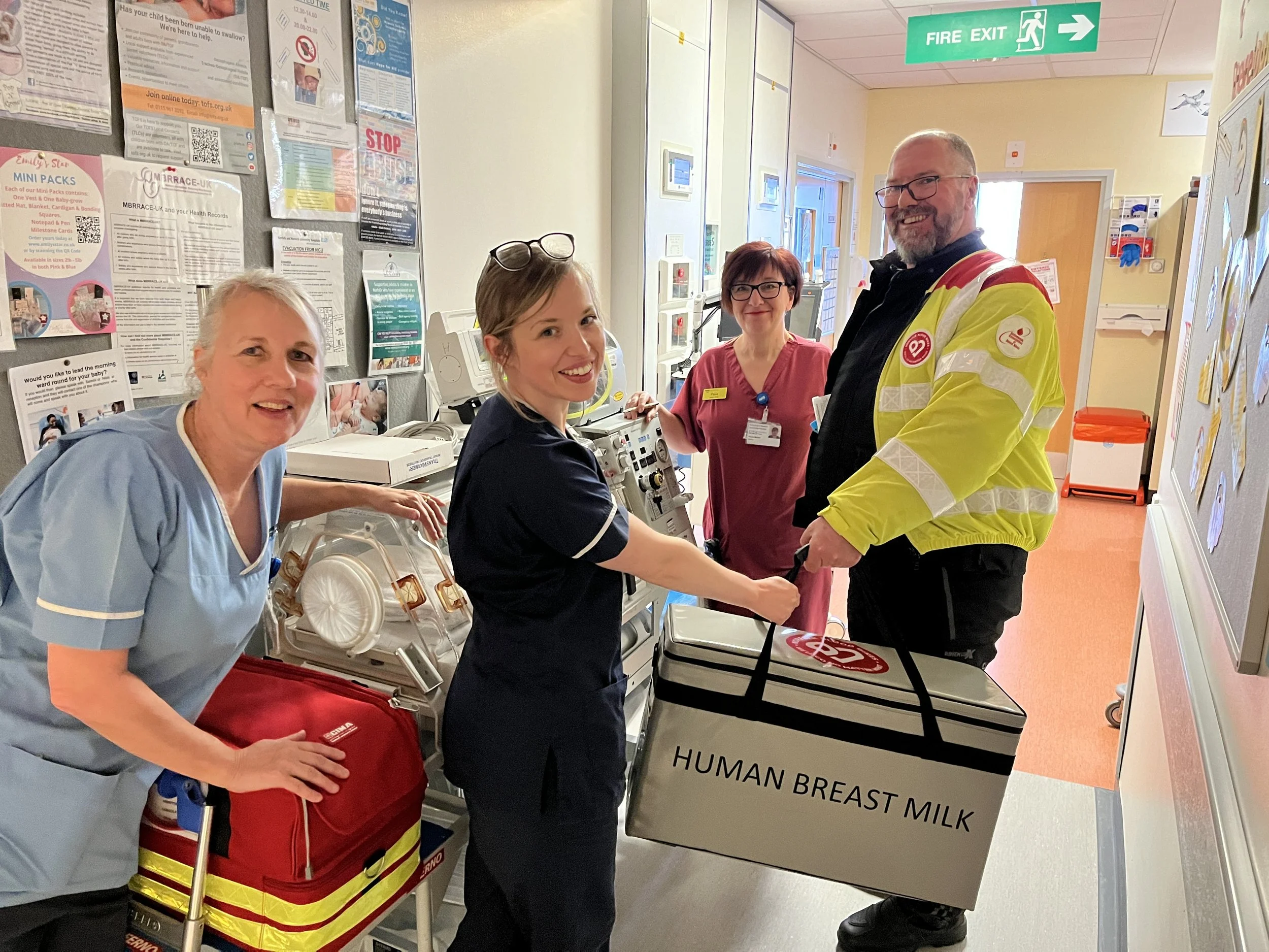 Medical professionals and a man holding a large donation box labeled 'Human Breast Milk' in a hospital corridor.