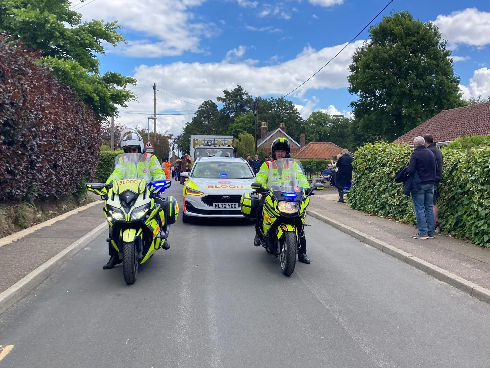 Two blood bikes in front of a blood donation vehicle, with onlookers on the sidewalk and a residential street with trees and houses in the background.
