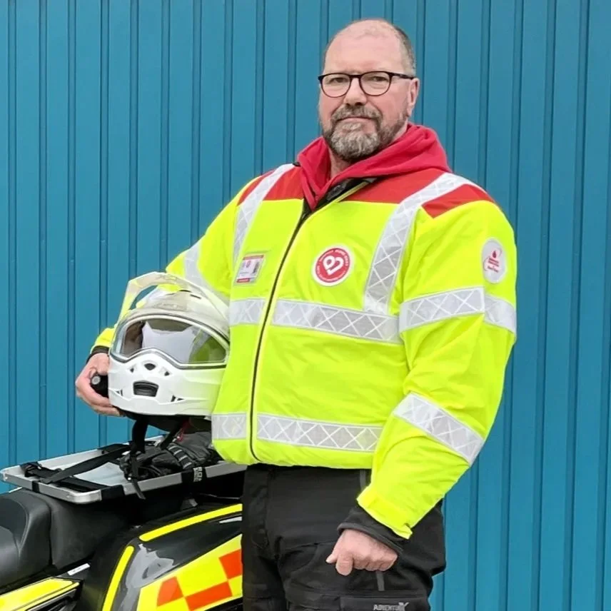 A volunteer in a high-visibility safety jacket holding a white helmet, standing next to a motorcycle with a blue metal wall in the background.