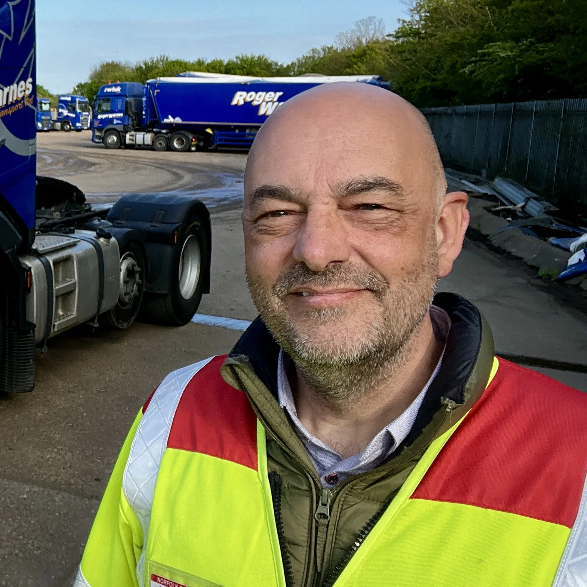 Man wearing a high-visibility yellow and red vest with trucks in the background.