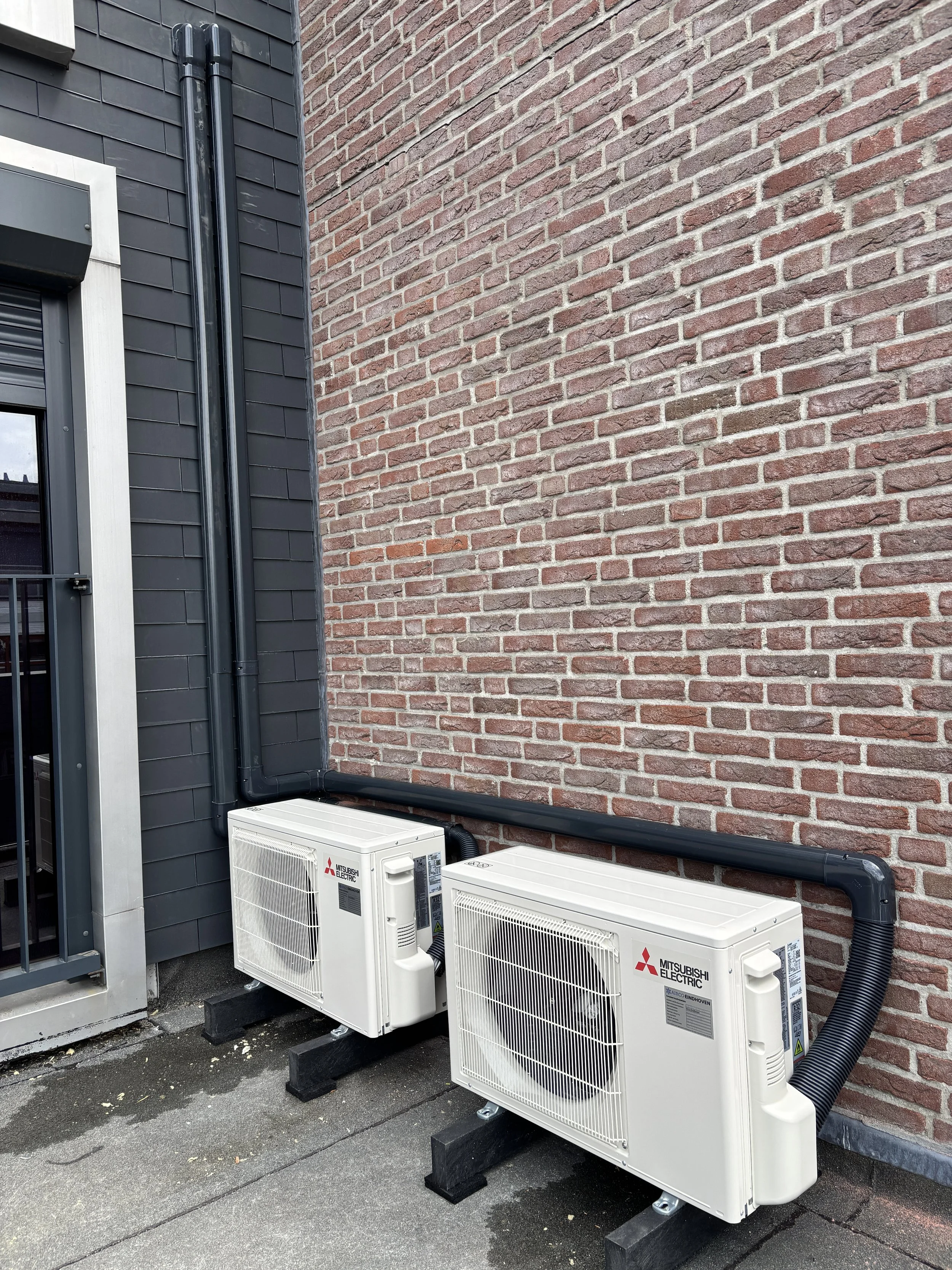 Twee Mitsubishi Electric airconditioning units buiten naast een zwartgepleisterde muur met een bakstenen muur ernaast.