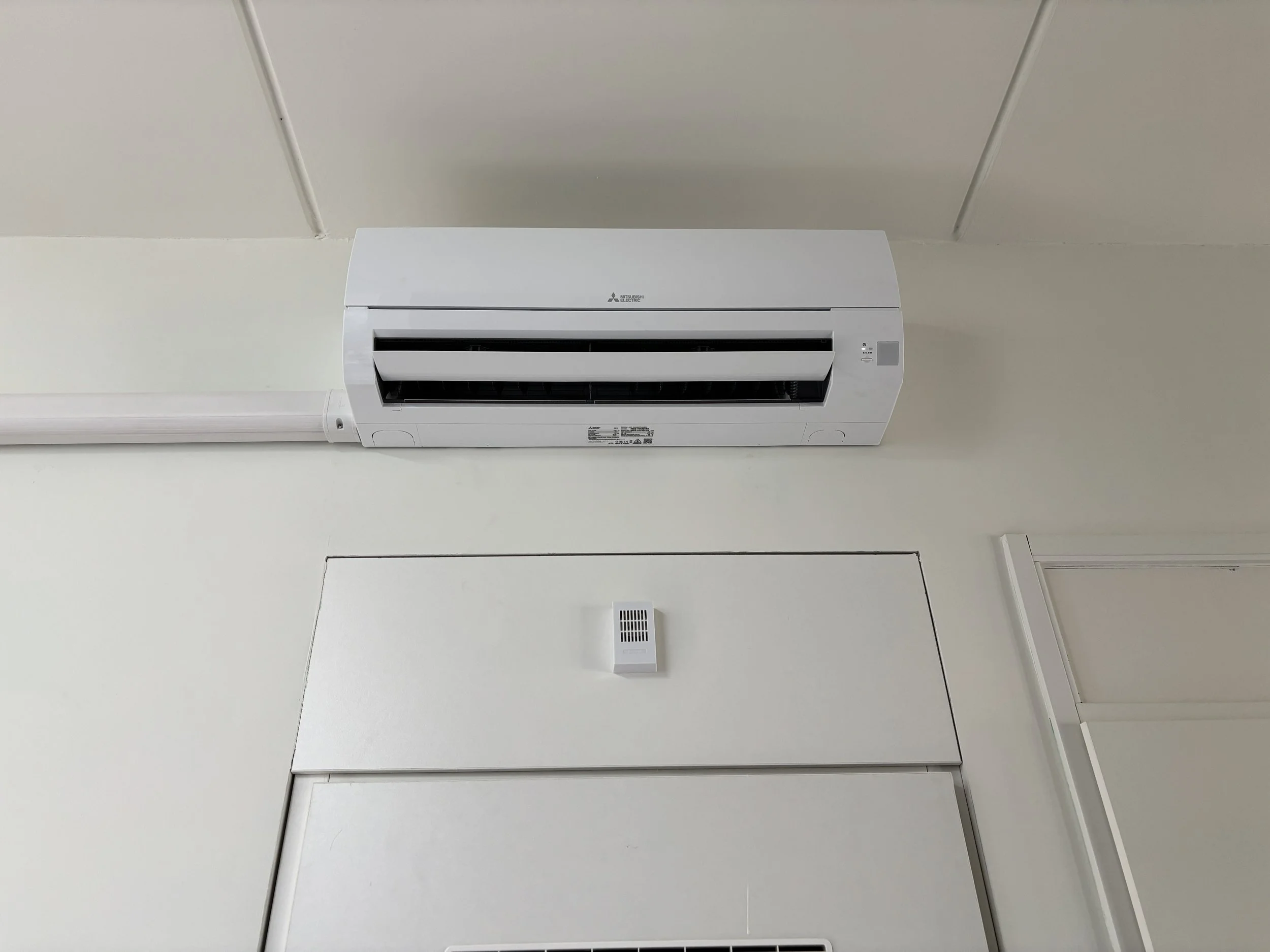 Witte muur gemonteerd met een airconditioner en een kleine ventilatie. Kleine onderdeeltjes en een witte leiding zichtbaar onder de airconditioner.
