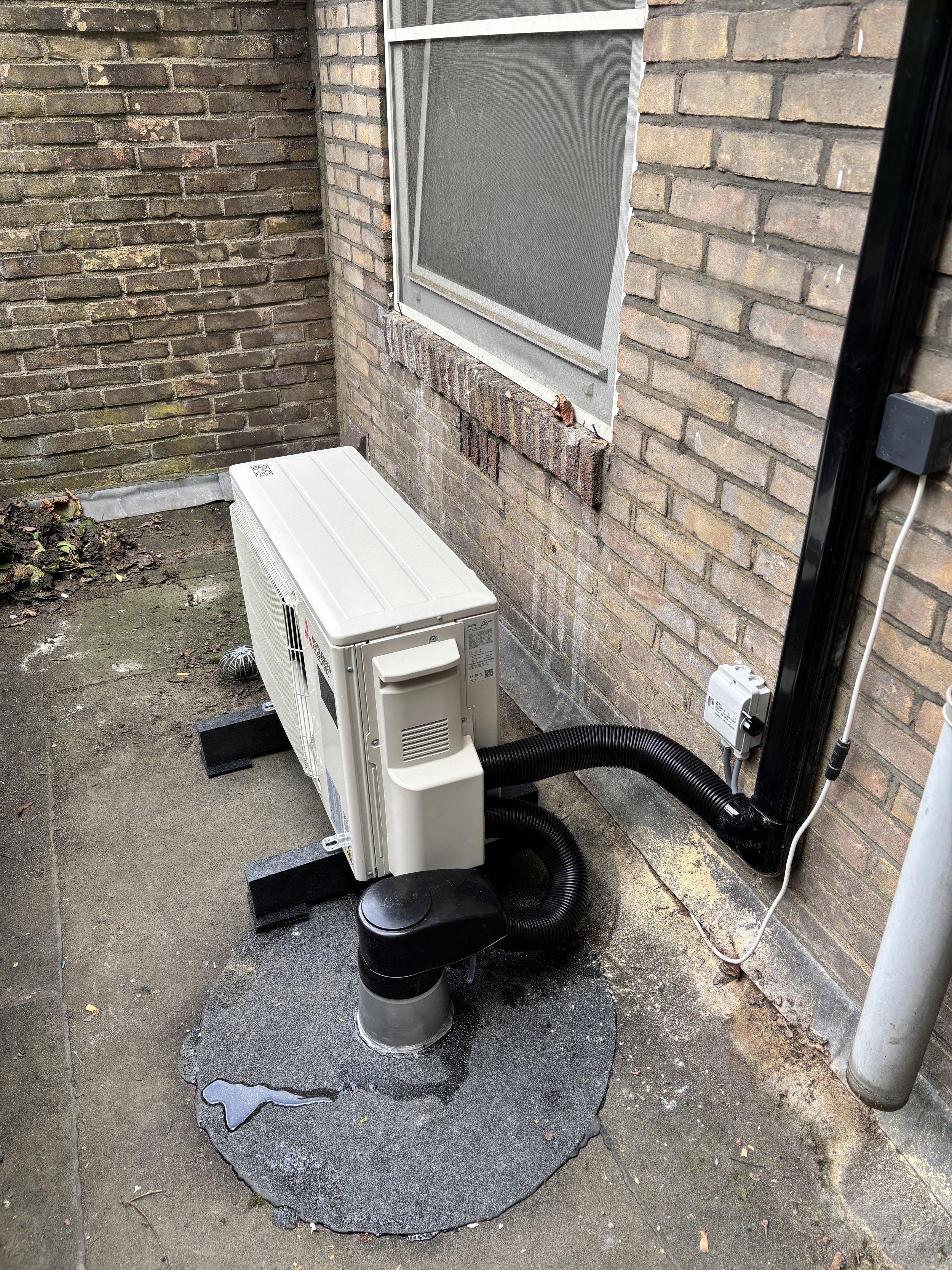 Een buitenunit van een airconditioningsysteem naast een bakstenen muur met een raam en elektra aansluitingen.