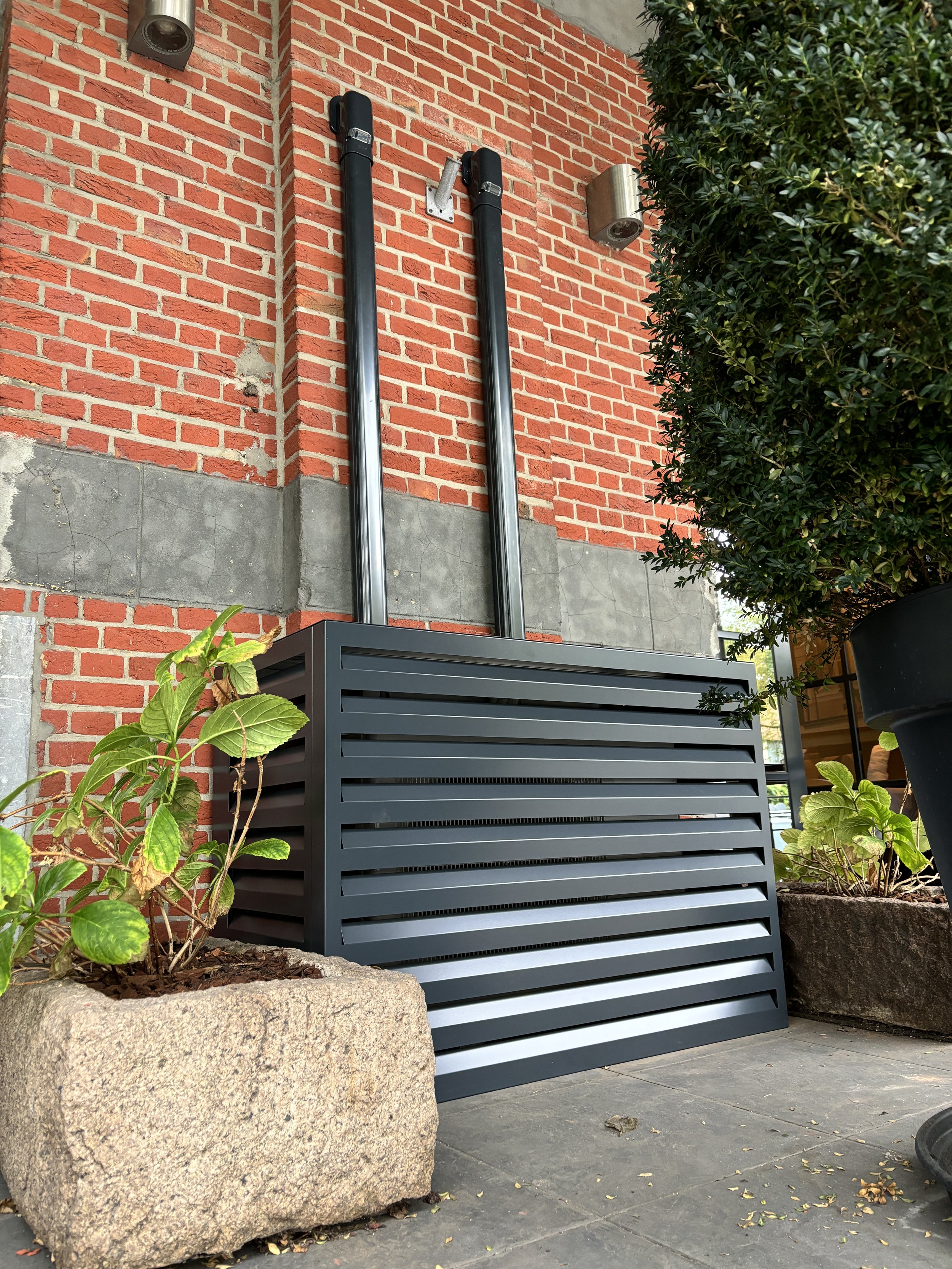 Modern grijze ventilatieplaat geplaatst in een stenen buitenmuur, omgeven door plantenbakken met groene planten en grote onkruid, met een rode bakstenen muur en buitenverlichting