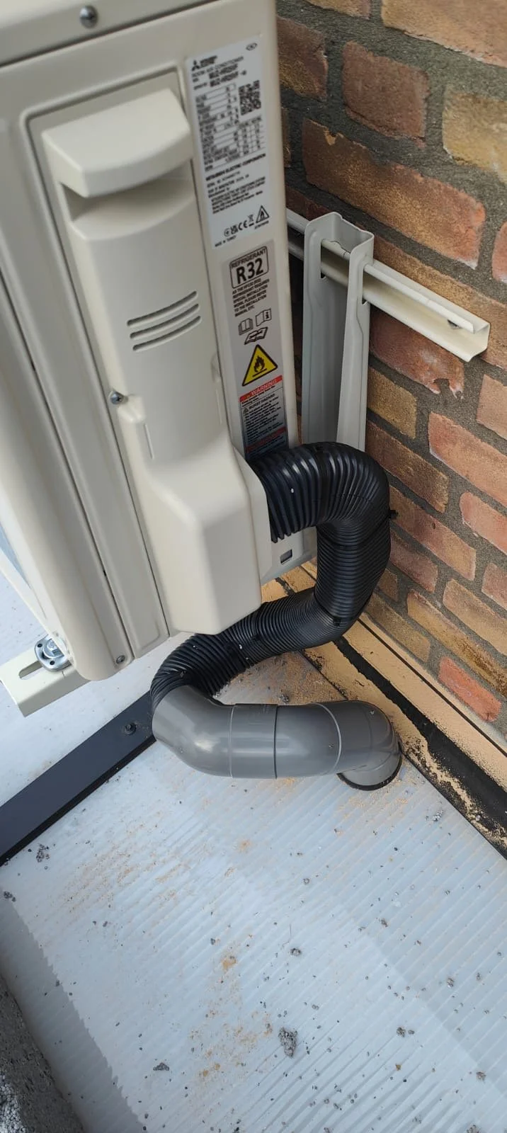 Elektrisch kastje met ventilatie en een afvoerslang tegen een bakstenen muur.