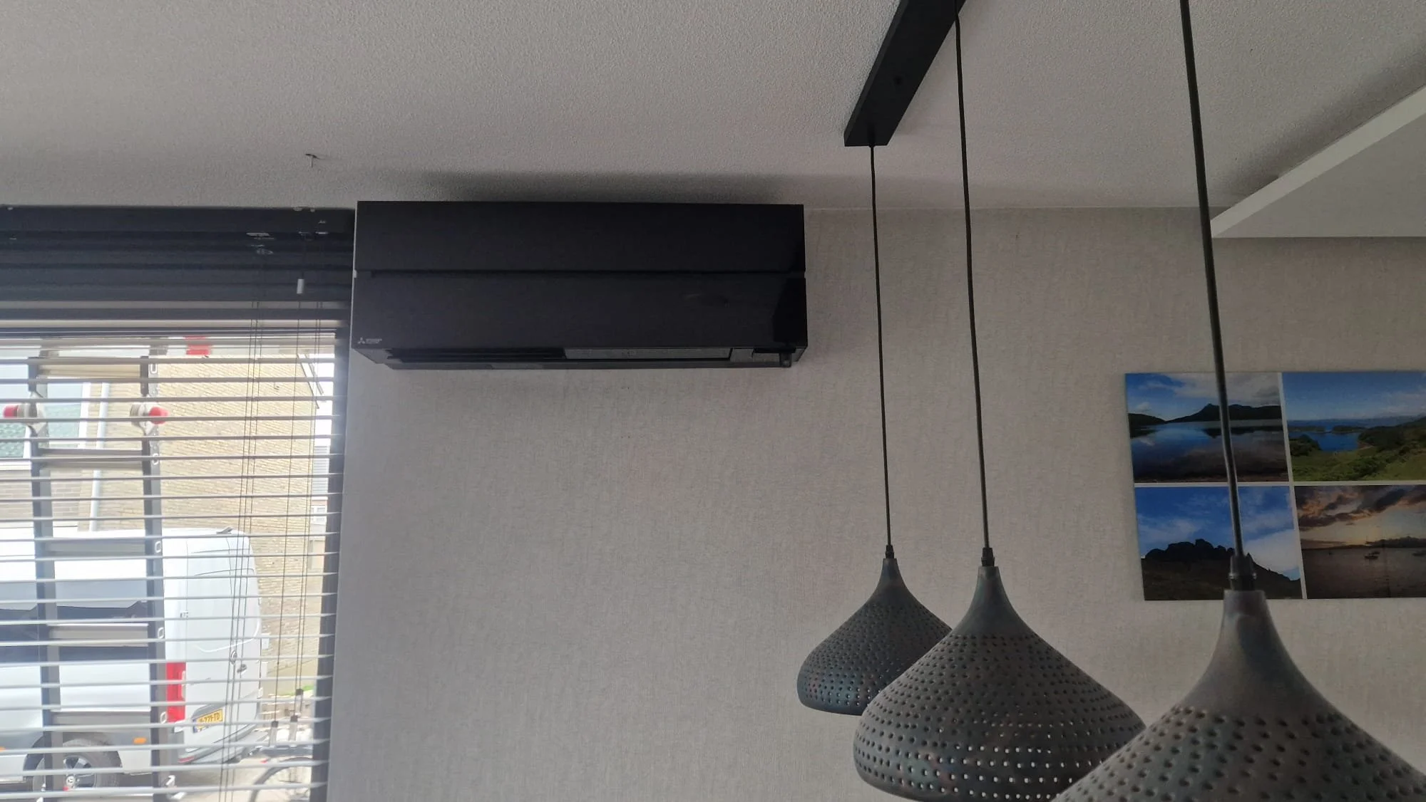 Een zwart airconditioner aan een witte wand, onder een raam met horizontale jaloezieën, met een foto van een landschap met water, bergen en een blauwe lucht op de muur.