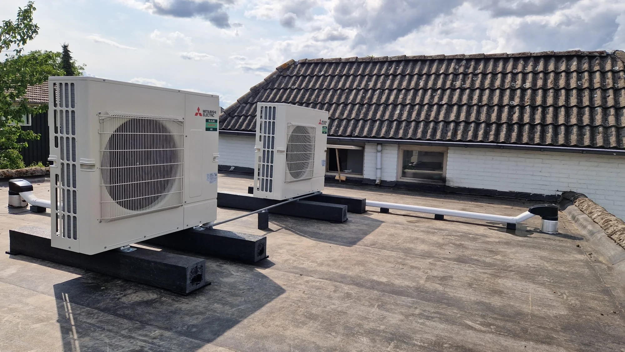 Twee Mitsubishi Electric airconditioning units op een plat dak, verbonden door witte leidingen, met een dakr ambientie en een huis met een rieten dak op achtergrond.