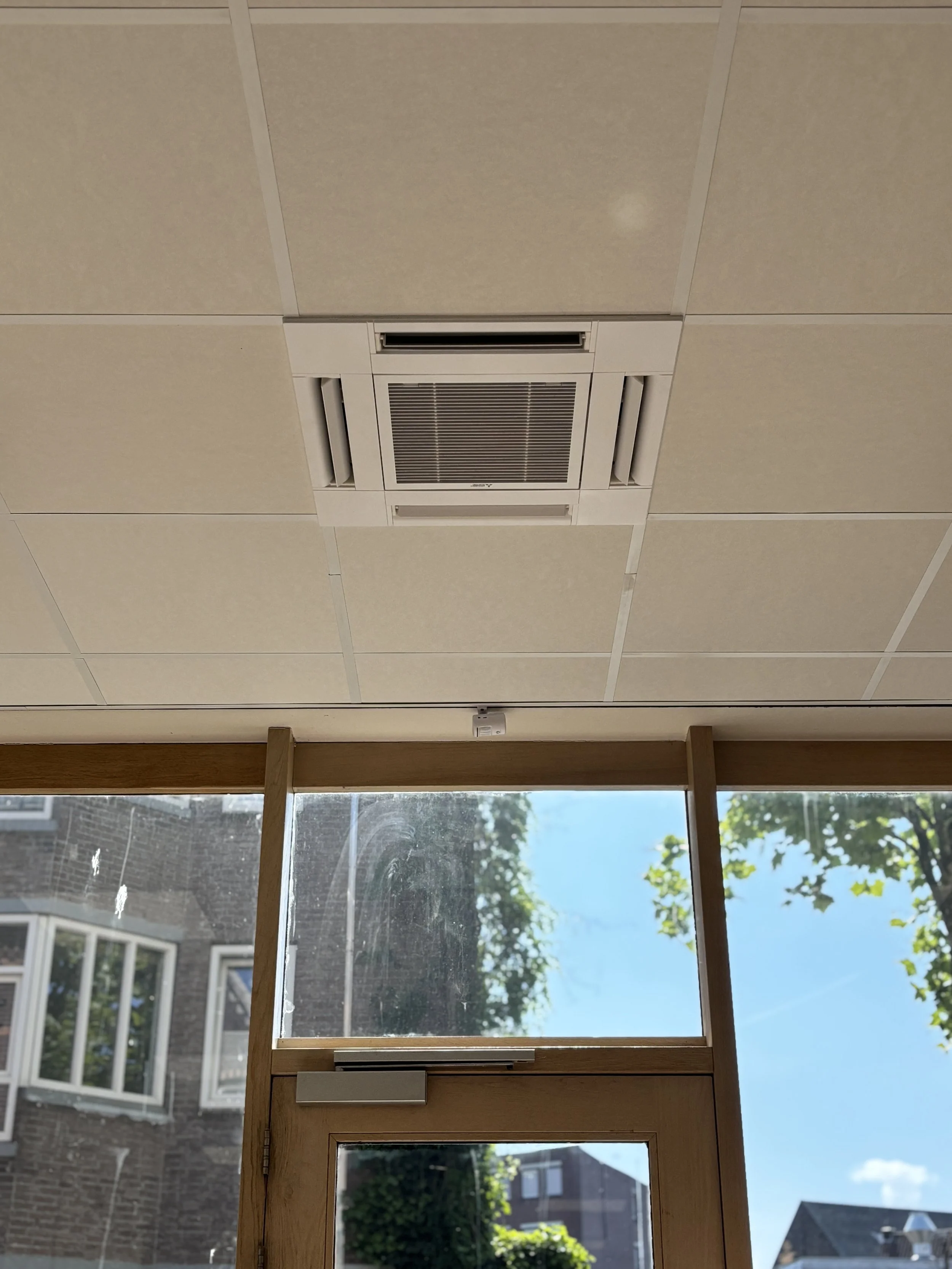 Binnenkant van een gebouw met een plafond met een ingebouwde airconditioning en grote ramen die uitzicht bieden op een blauwe luchten, bomen en gebouwen buiten.