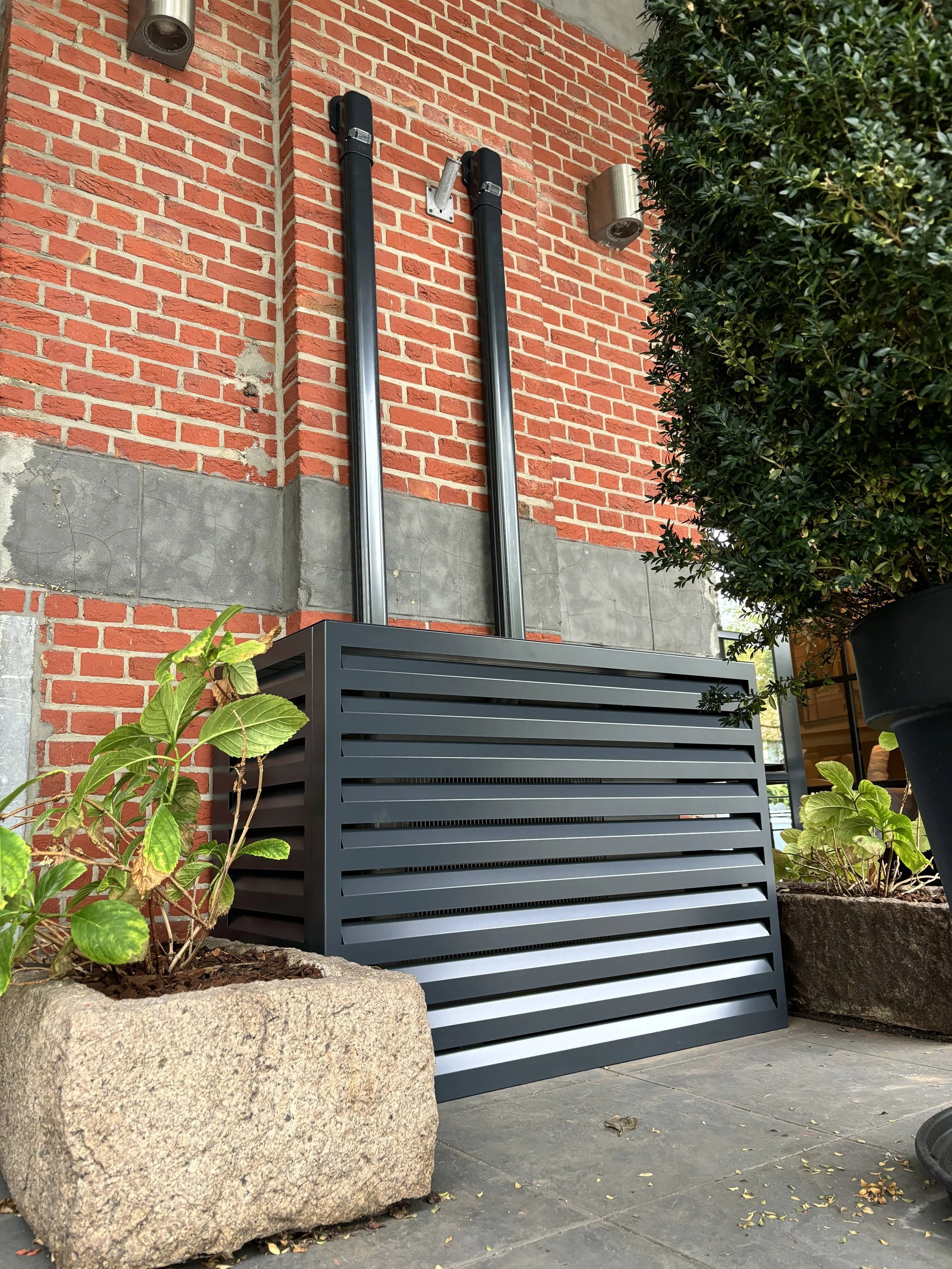 Een grijze ventilatorombouw op een tegeldopleiding naast planten in stenen bakken, tegen een rode bakstenen muur met groene struiken ernaast.