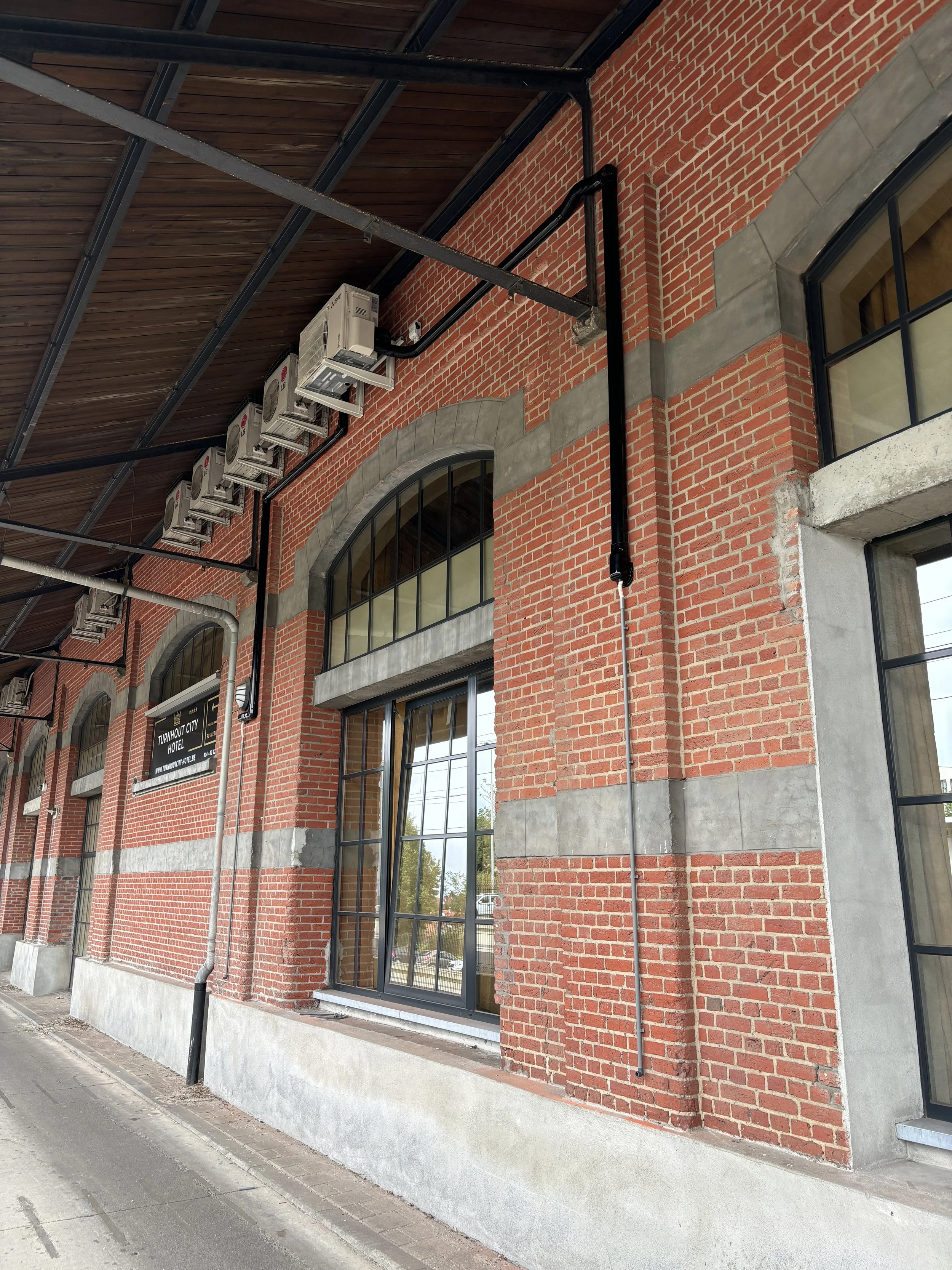 Lijn met meerdere airconditioners en afvoersystemen langs een rode bakstenen muur van een gebouw met grote ramen.