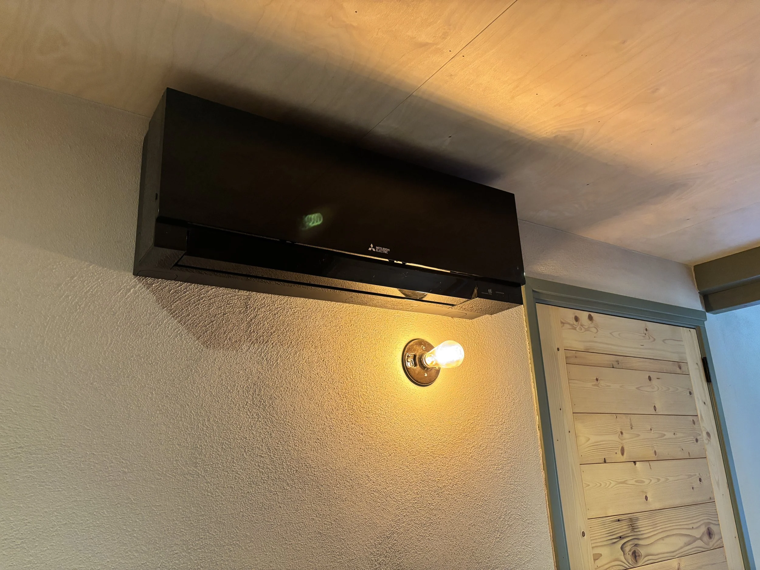 Een zwart airconditioner aan de muur onder een verlichte plafondlamp in een kamer met houten wandpanelen.