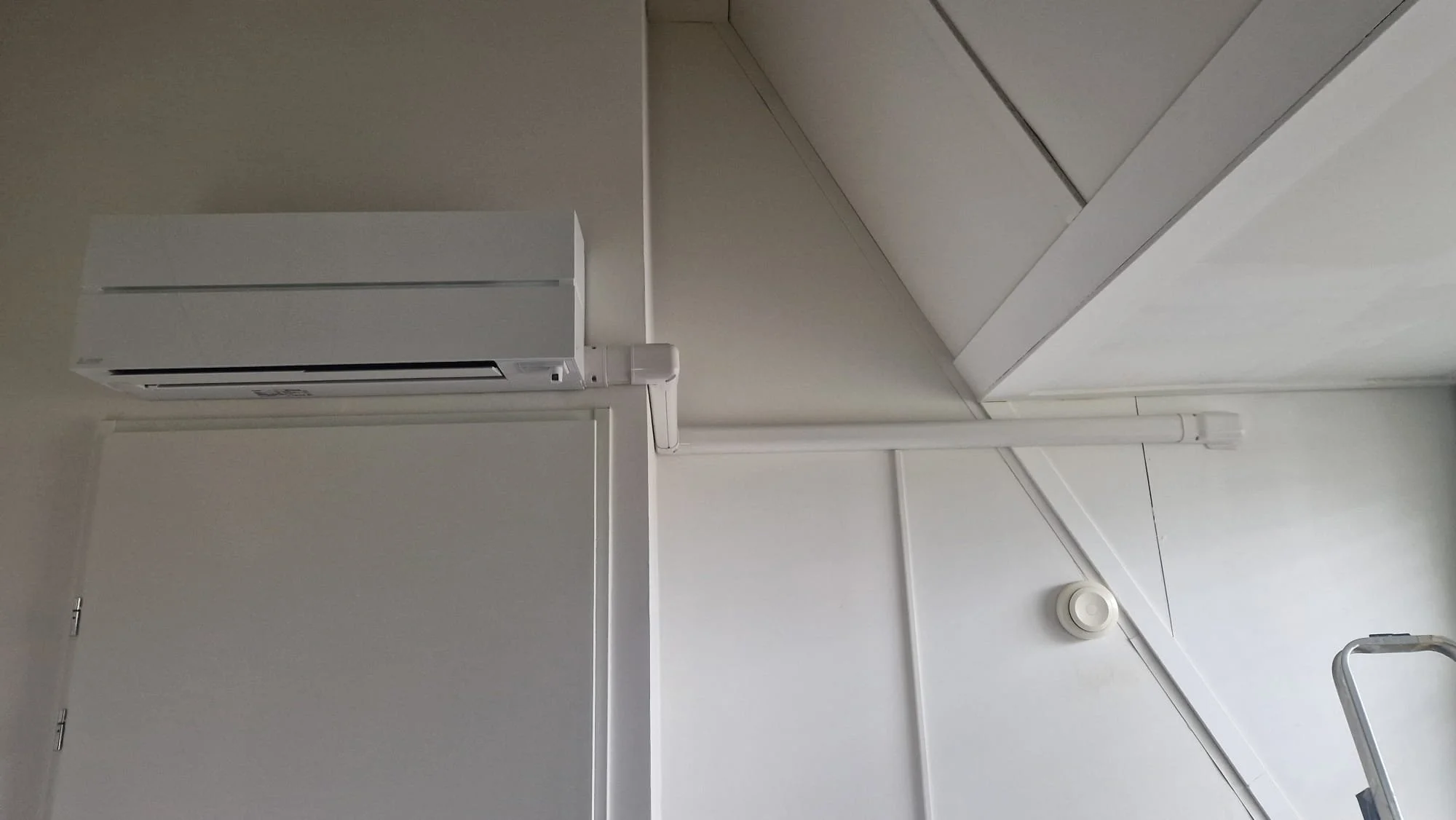 Binnenkant van een kamer met een witte airconditioner en een witte muur. There is een lichtpunt op het plafond, en een ladder is gedeeltelijk zichtbaar aan de rechterkant.