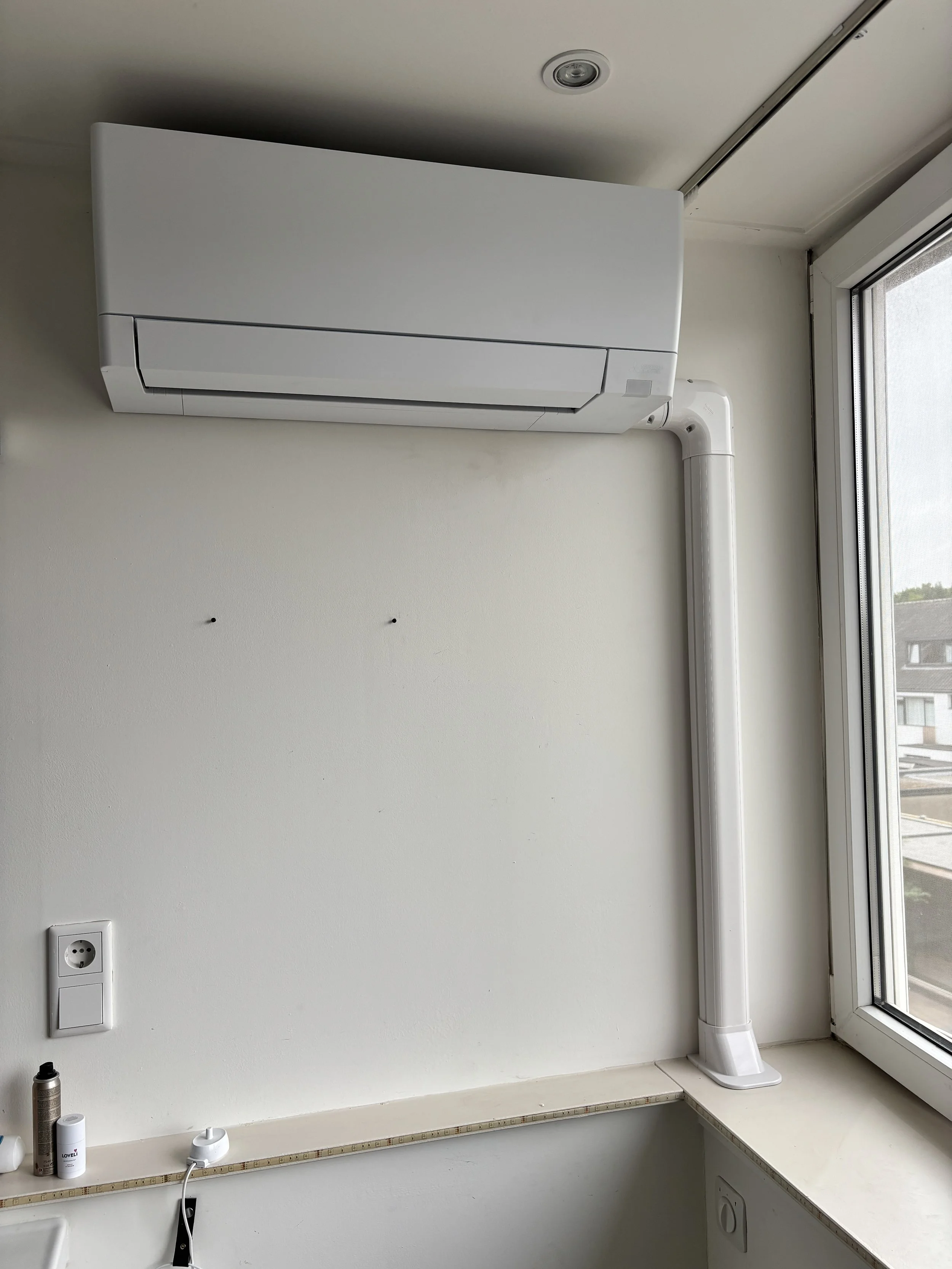 Een witte airconditioner is bevestigd aan de muur in een kamer met een raam dat uitkijkt op andere gebouwen buiten.