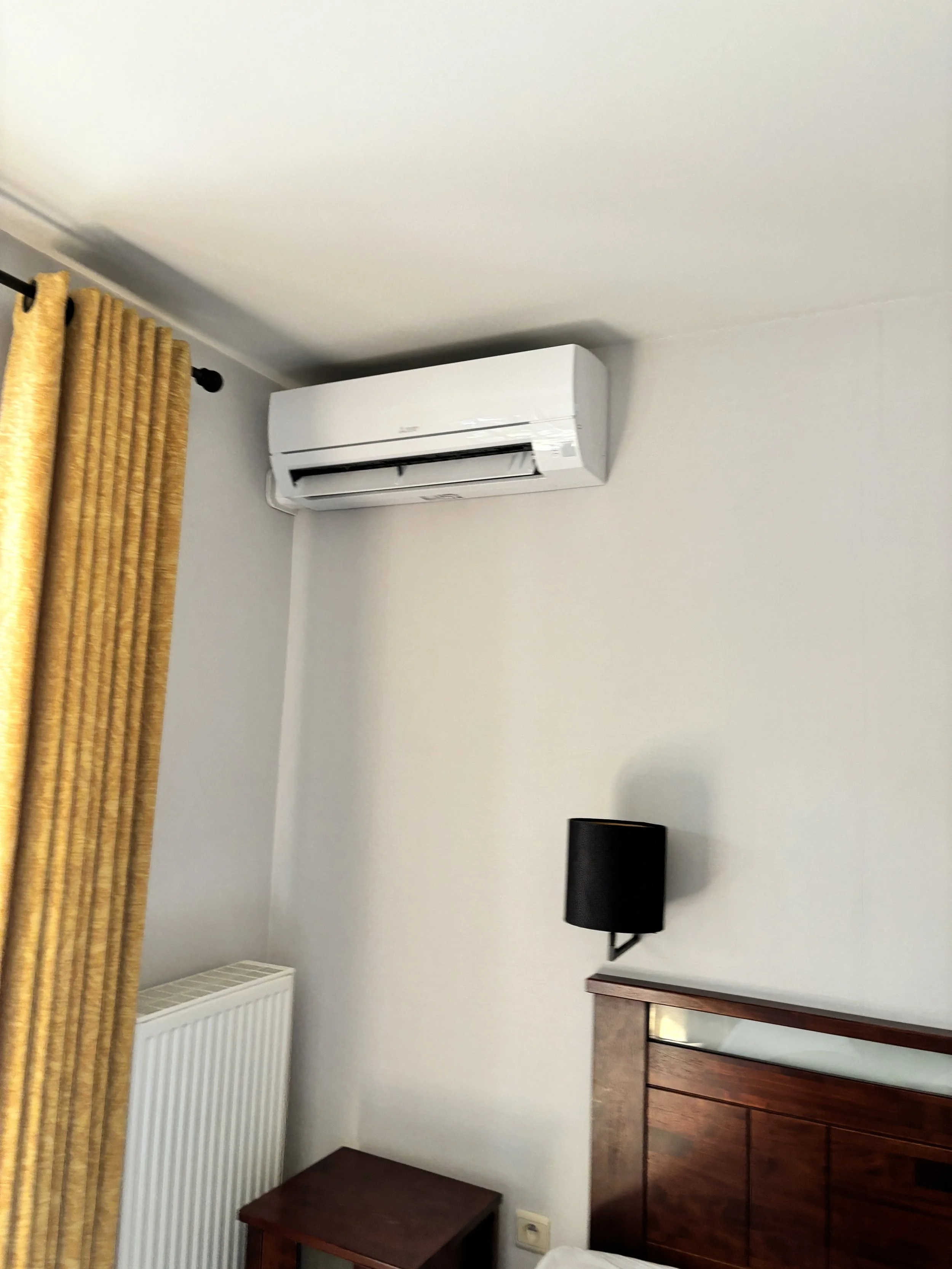 Een kamer met witte muren, een airconditioner aan het plafond, een zwarte muurmontage lamp, een radiateur, een bruine houten kast en gele gordijnen.