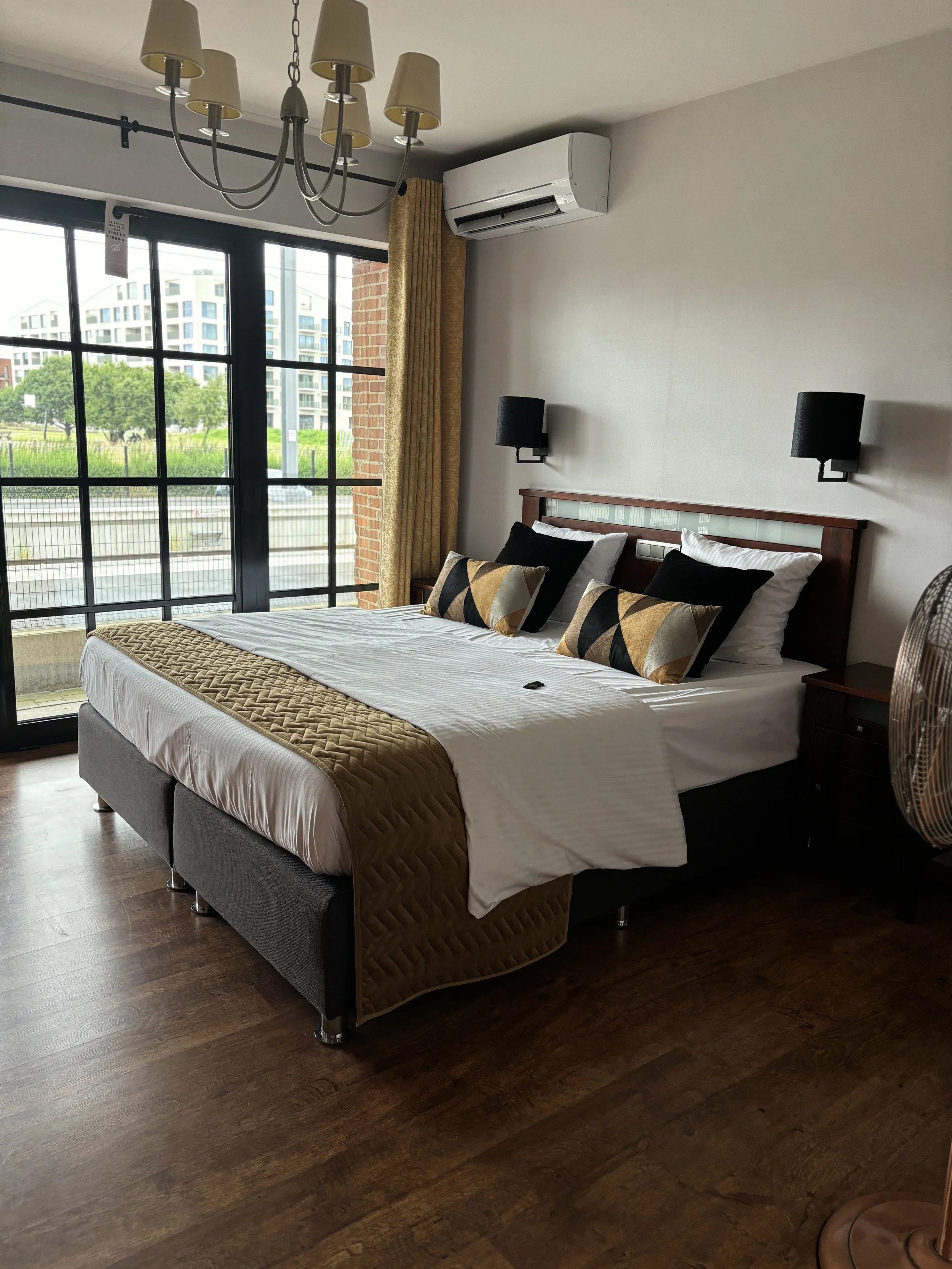 Een hotelkamer met een groot bed, kussen, en dekbedovertrek in zwart, wit, en goudkleurige patronen. Er zijn grote ramen met uitzicht op een modern gebouwencomplex en groene bomen. Er hangt een plafondlamp en er zijn muurhanglampen. Een airconditione