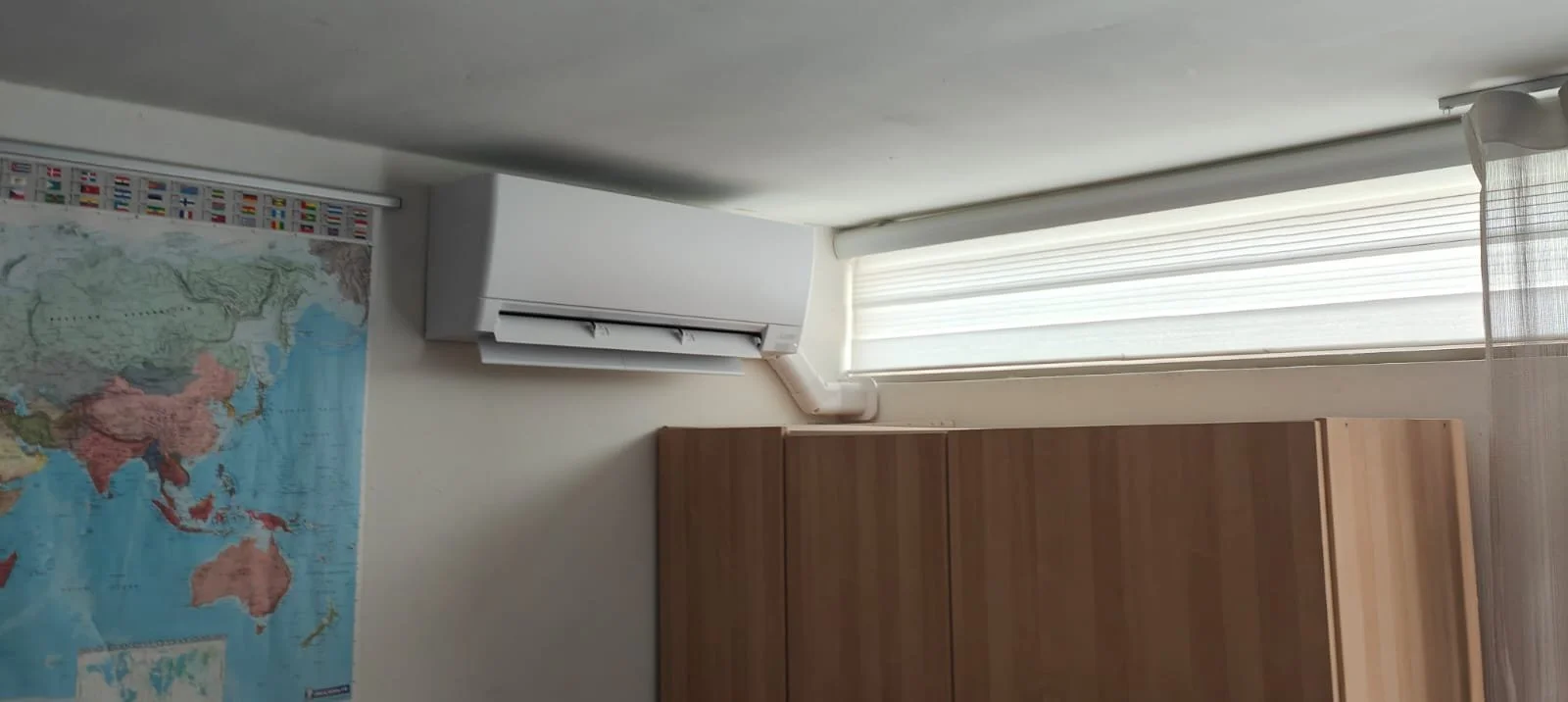 Binnenkant van een kamer met een wereldkaart op de muur, een luchtconditioner, een raam met jaloezieën en een houten kast.