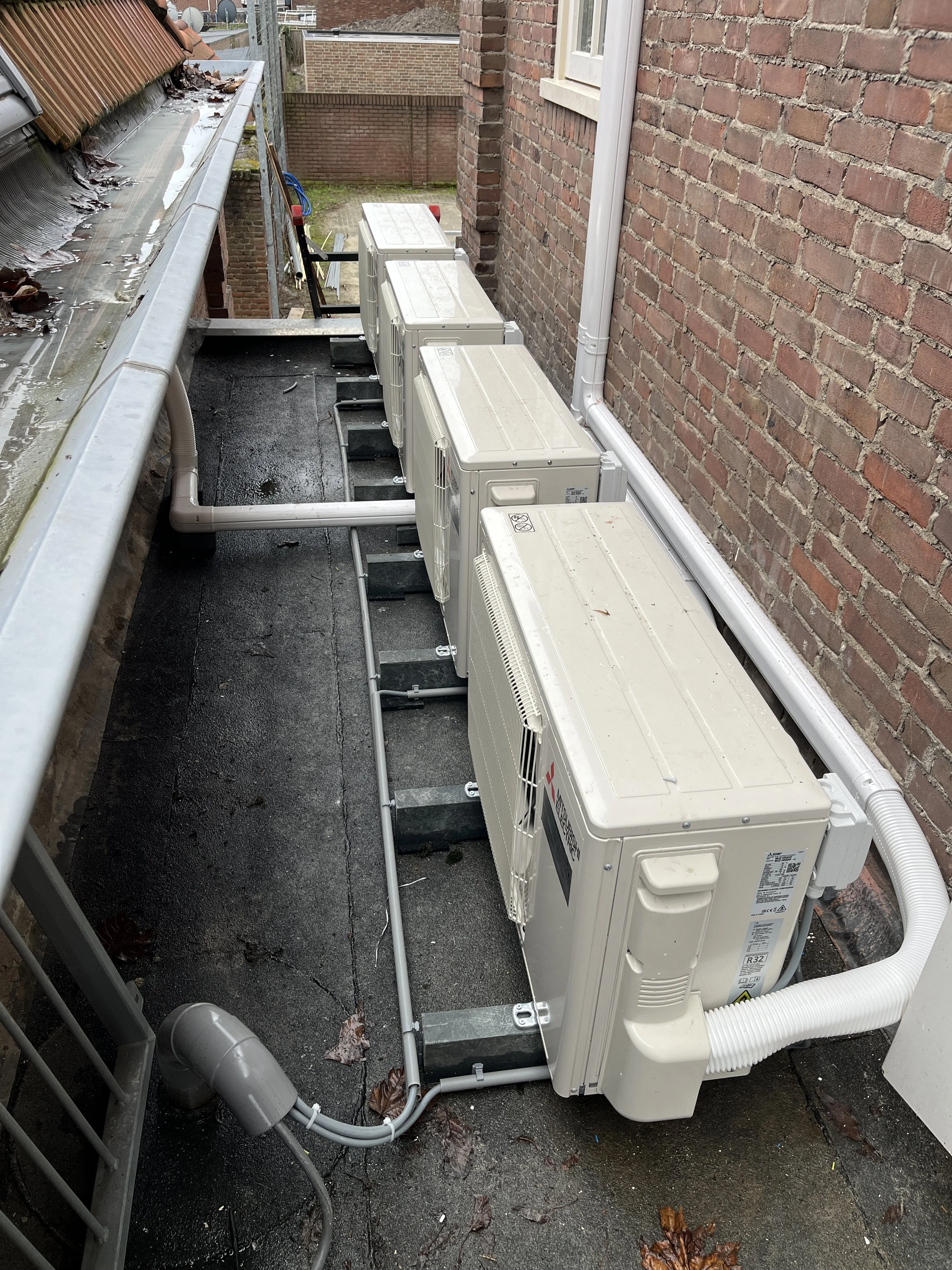 Rijen buitenunits van airconditioning of warmtepompen op een dak naast een bakstenen muur, met zicht op afvoeringsbuizen en ventilatoren.