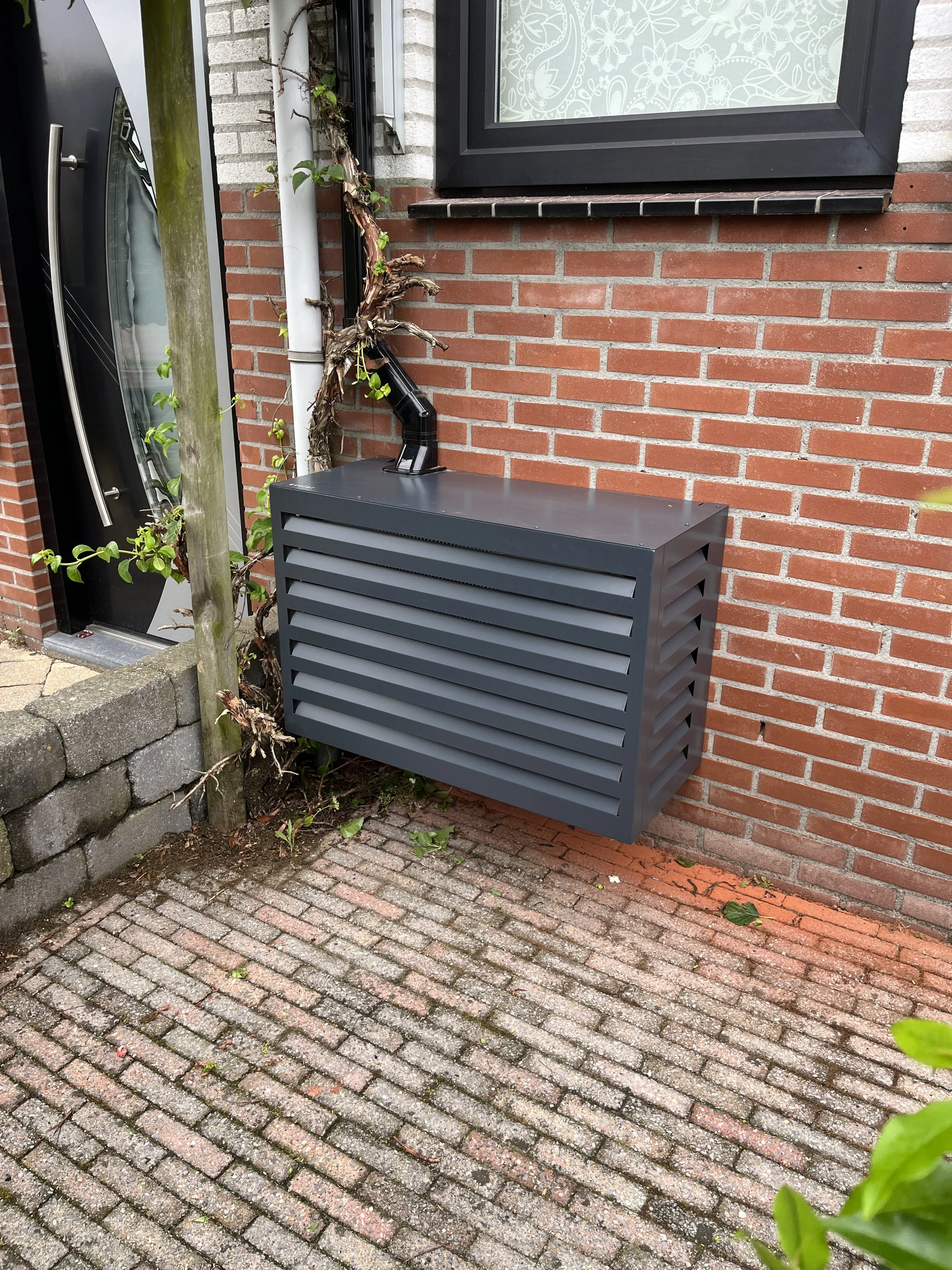 Een buitenlucht van een huis met rode bakstenen muur, een zwart rooster aan de muur, een zwarte radiator, een deur met een spiegel, en een raam met witte versieringen.