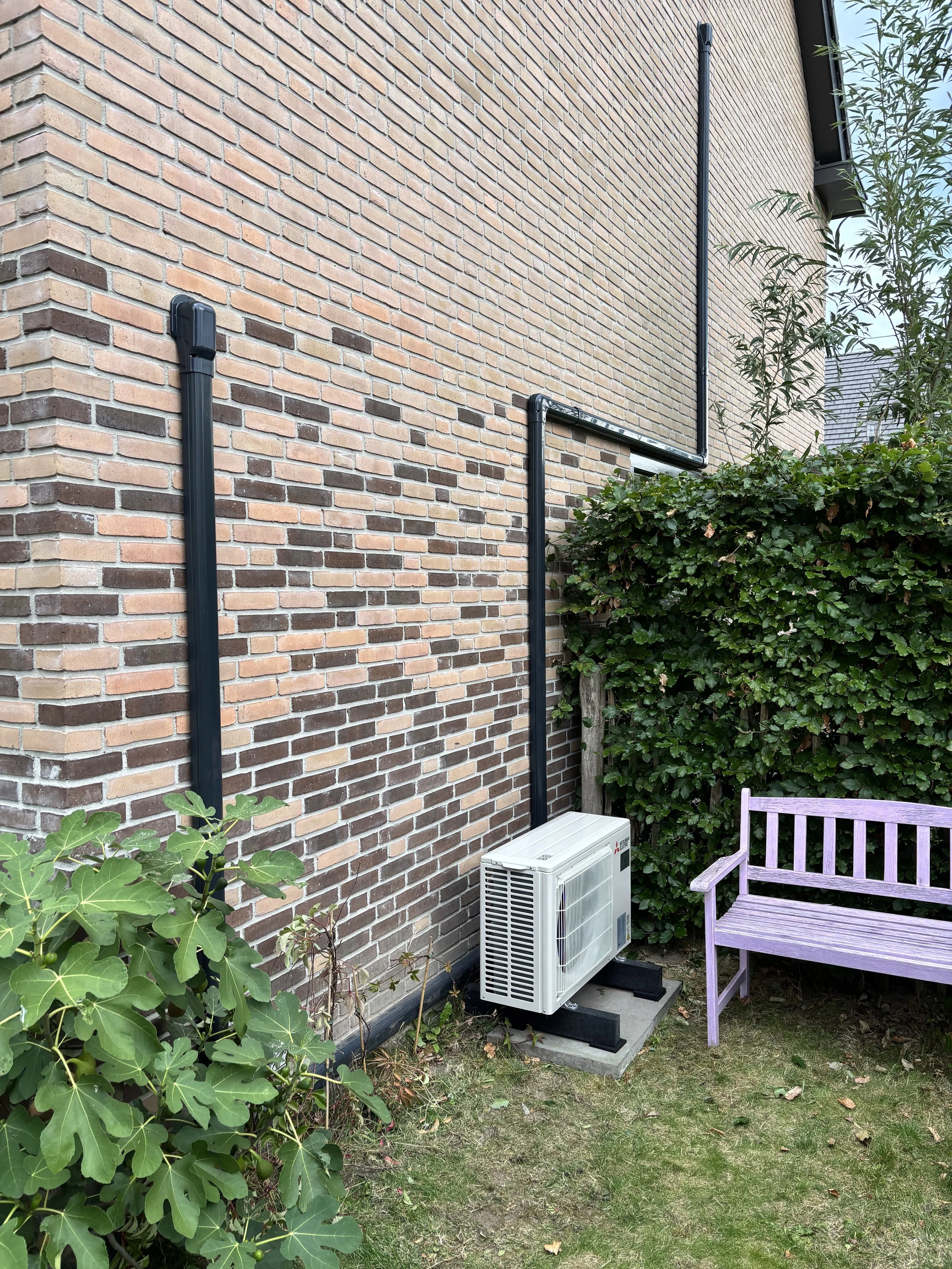 Een tuin met een bakstenen muur, een airconditioning-unit op een betonnen sokkel, een paar zwarte leidingen en een roze houten bankje.