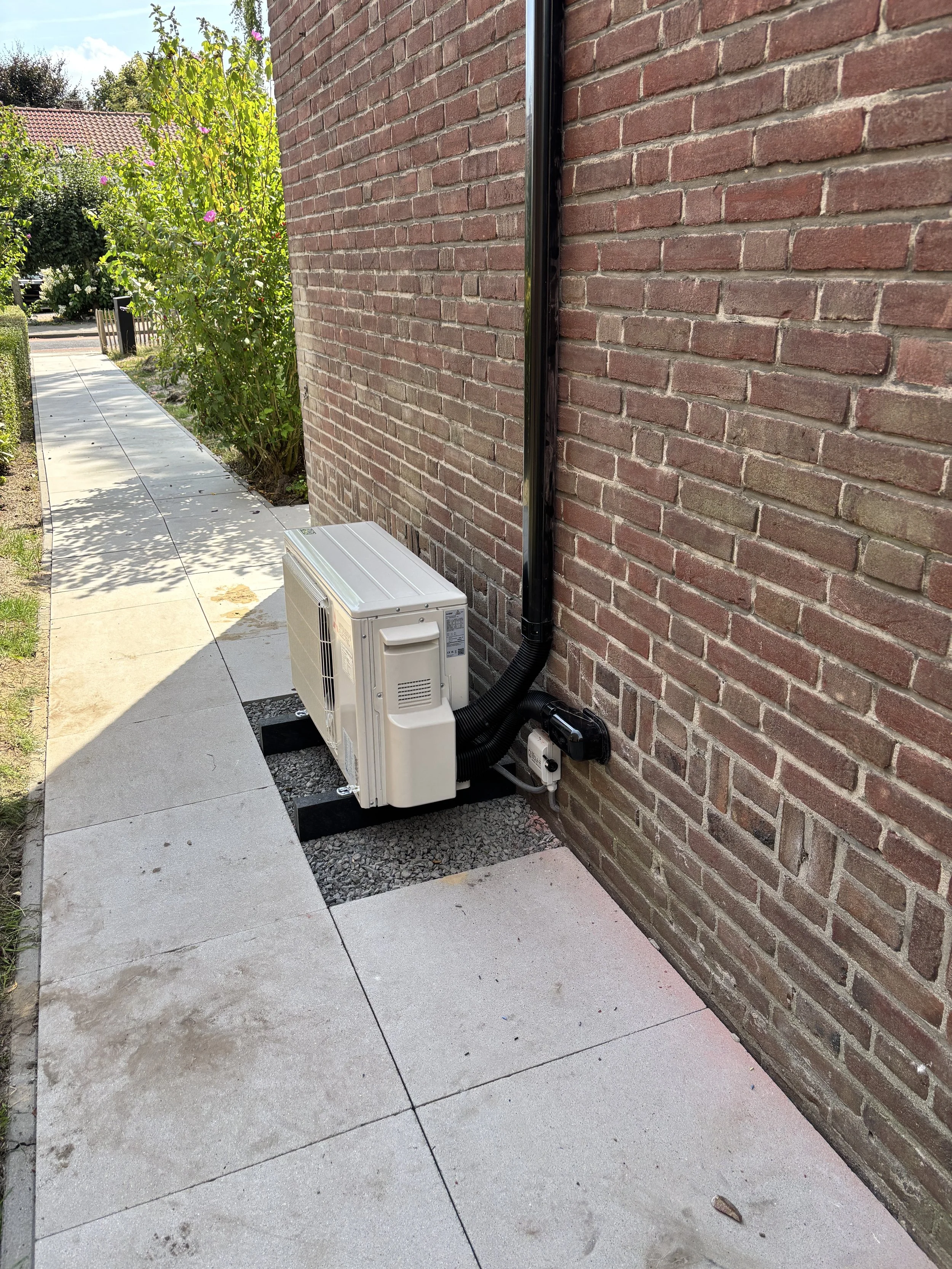 Een airconditioningsunit buiten naast een bakstenen muur van een gebouw, bevestigd op een betonnen ondergrond.