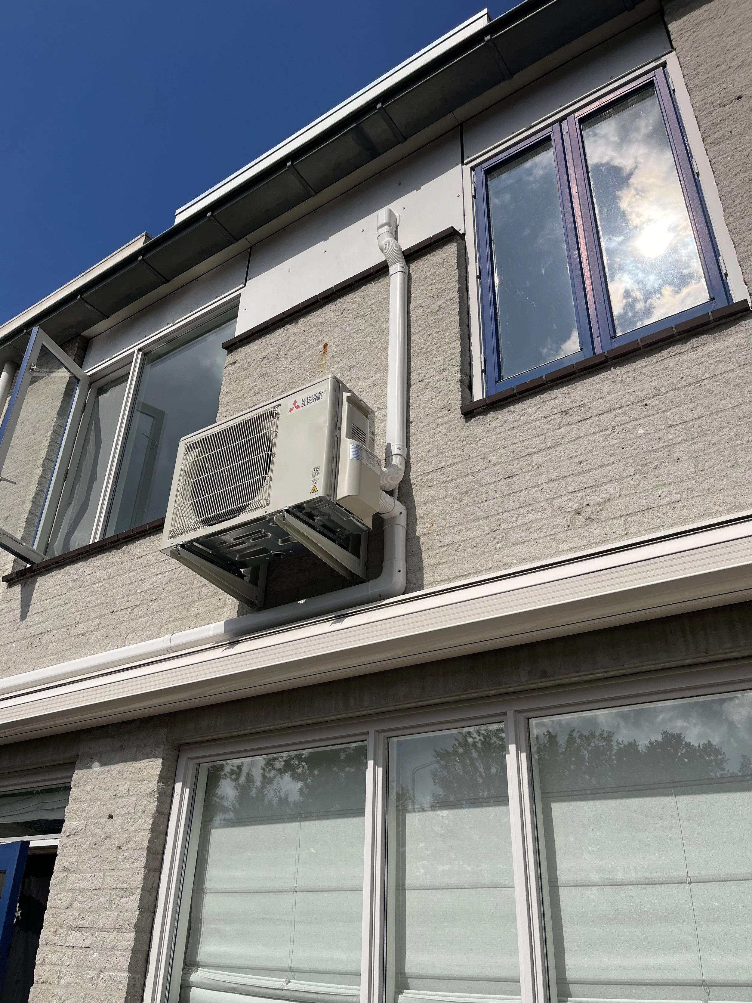 Uitzicht op de buitenmuur van een gebouw met een airconditioning en een raam weerspiegeld in het glas, onder een strakblauwe hemel.