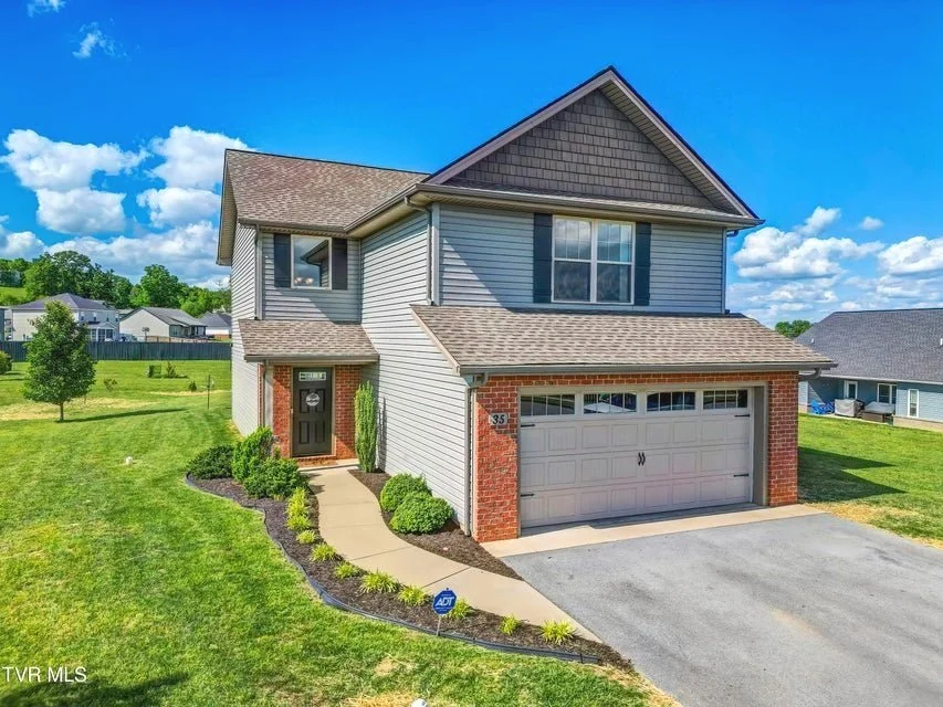 Two-story suburban house with gray siding, brick accents, and a gray garage door, surrounded by a green lawn and a clear blue sky.