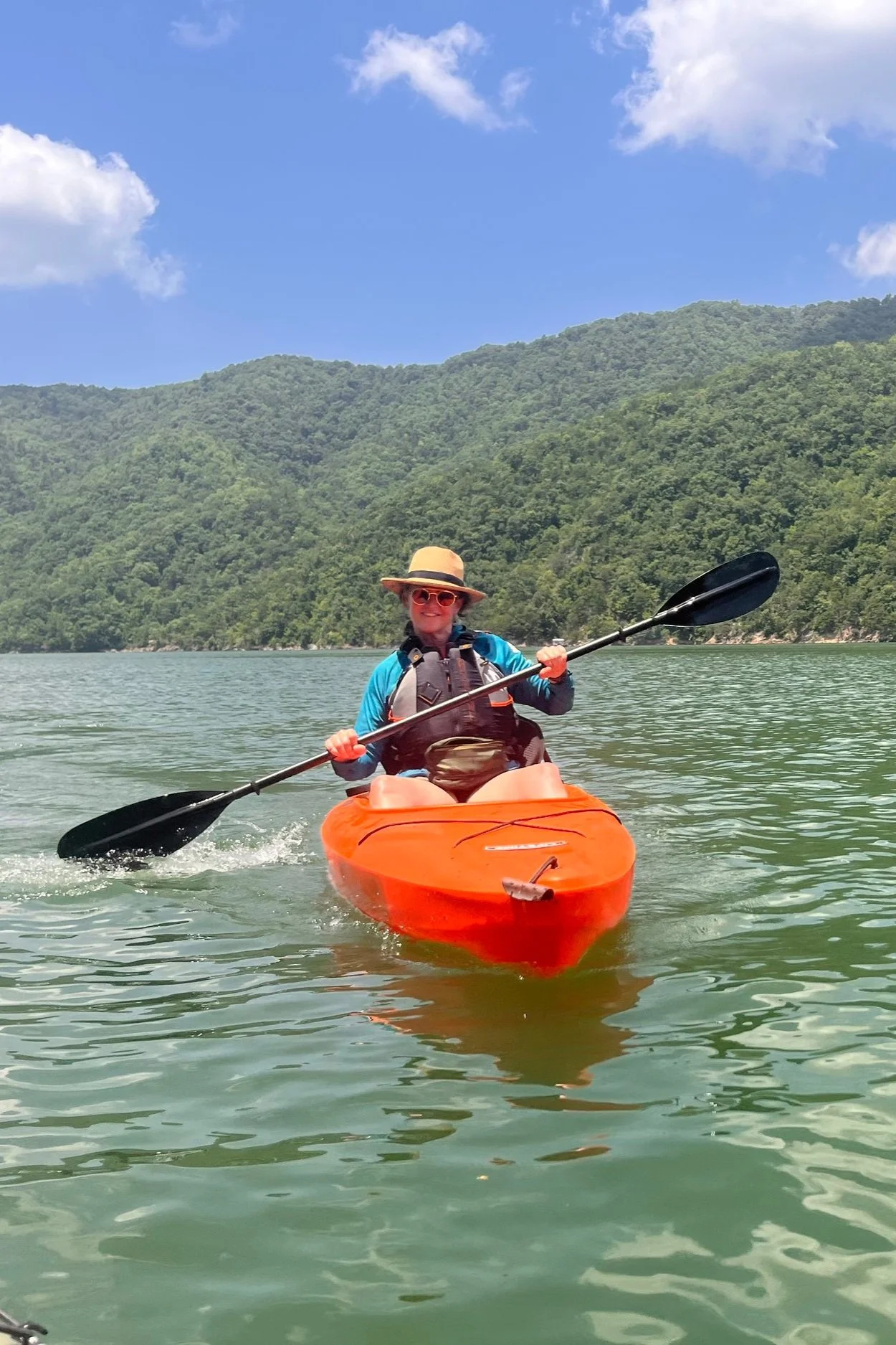 An orange kayak on Watauga Lake on a sunny day featuring a life vest and a smiling woman.